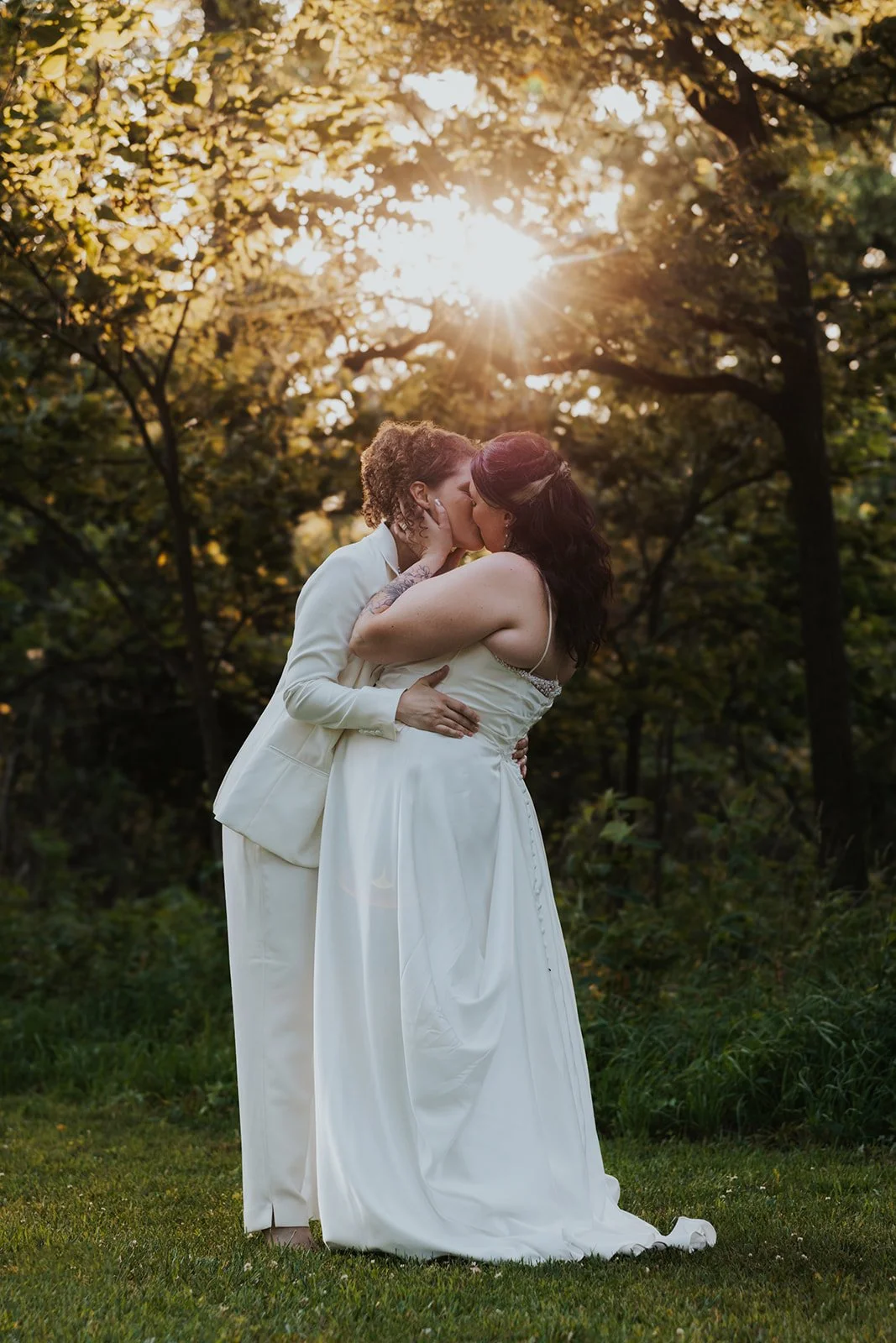 Two brides kissing each other closely with sunset behind them and lots of trees and greenery