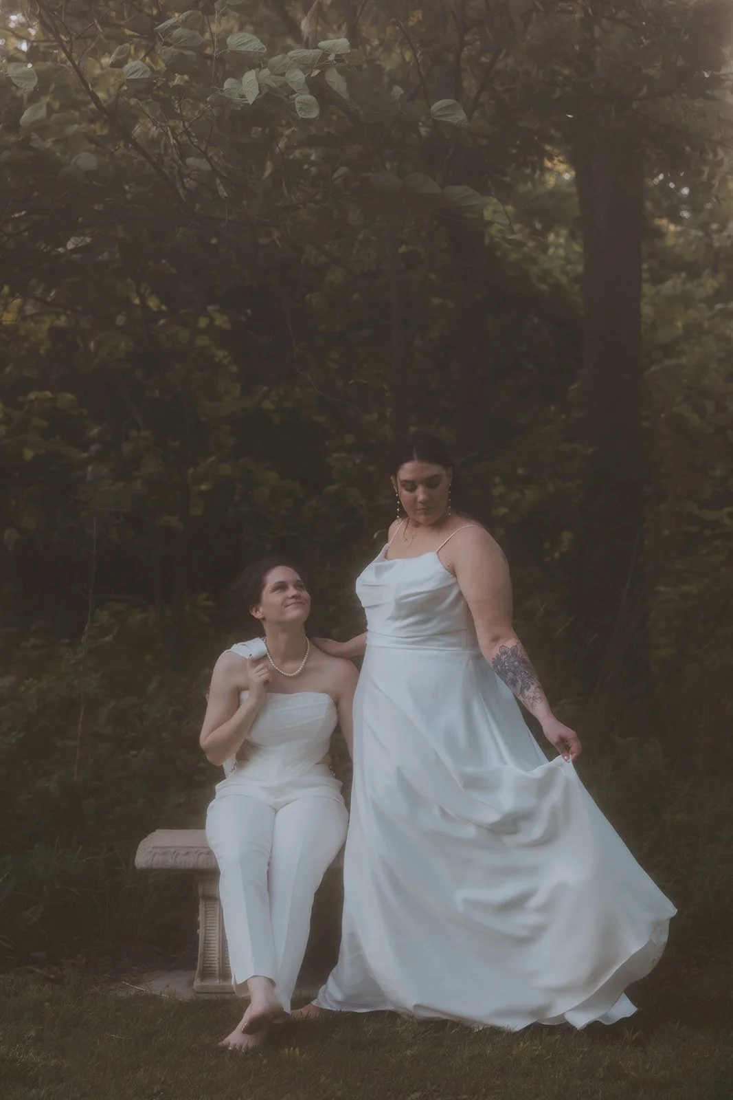 filmy, milky photo with bride in dress standing next to seated bride in suit looking up at her