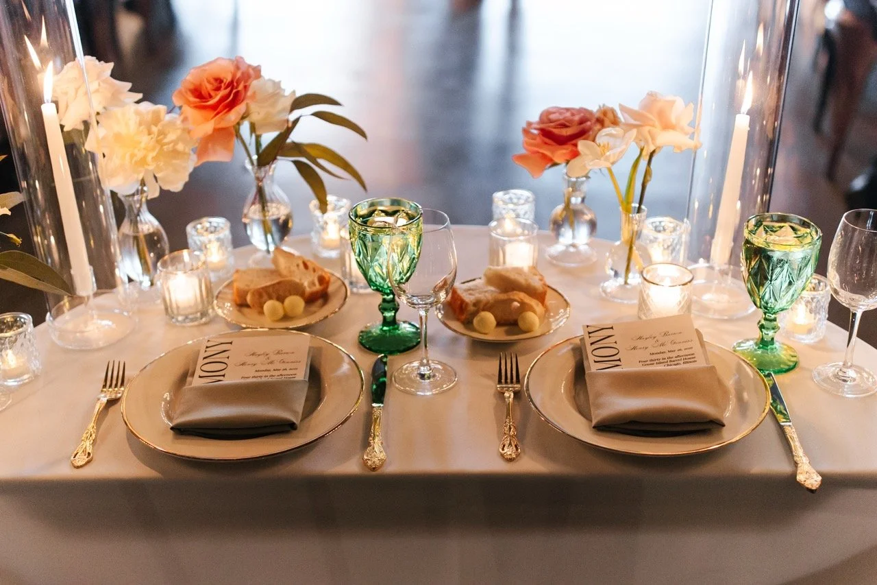 Sweetheart table setup with plates, menus, bud vases, candles, and green water goblets