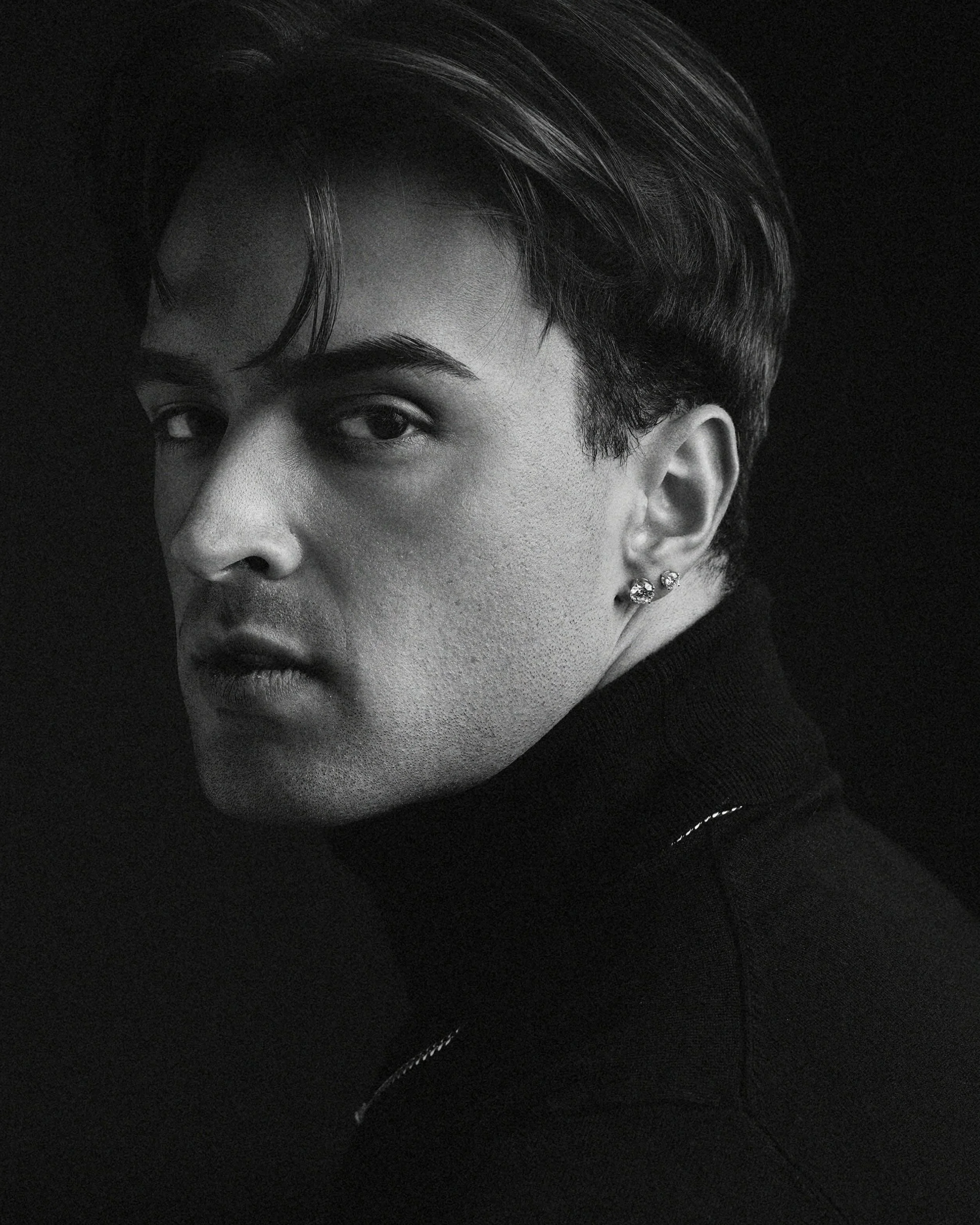 Black and white portrait of a young man with short, styled hair, wearing earrings and a turtleneck, looking over his shoulder against a dark background.
