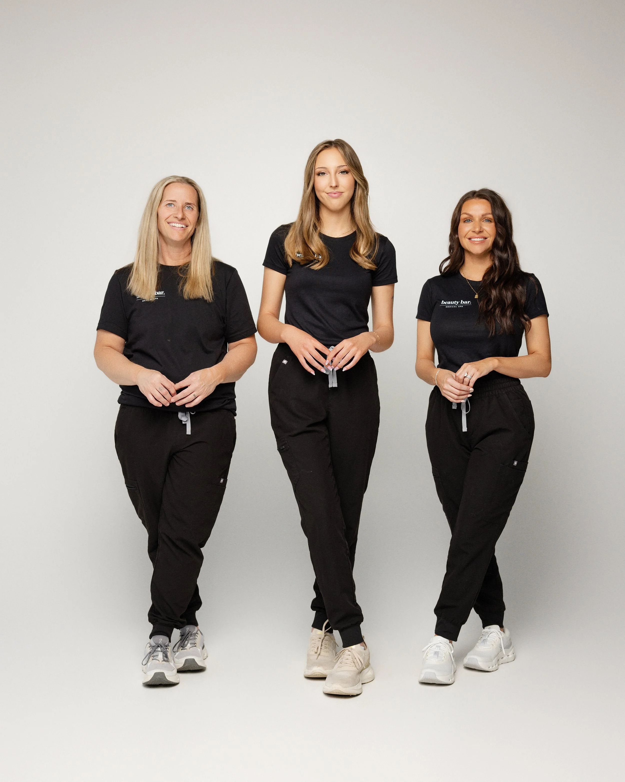 Three women standing together against a plain white wall, wearing black t-shirts and black pants with sneakers, smiling and looking at the camera.