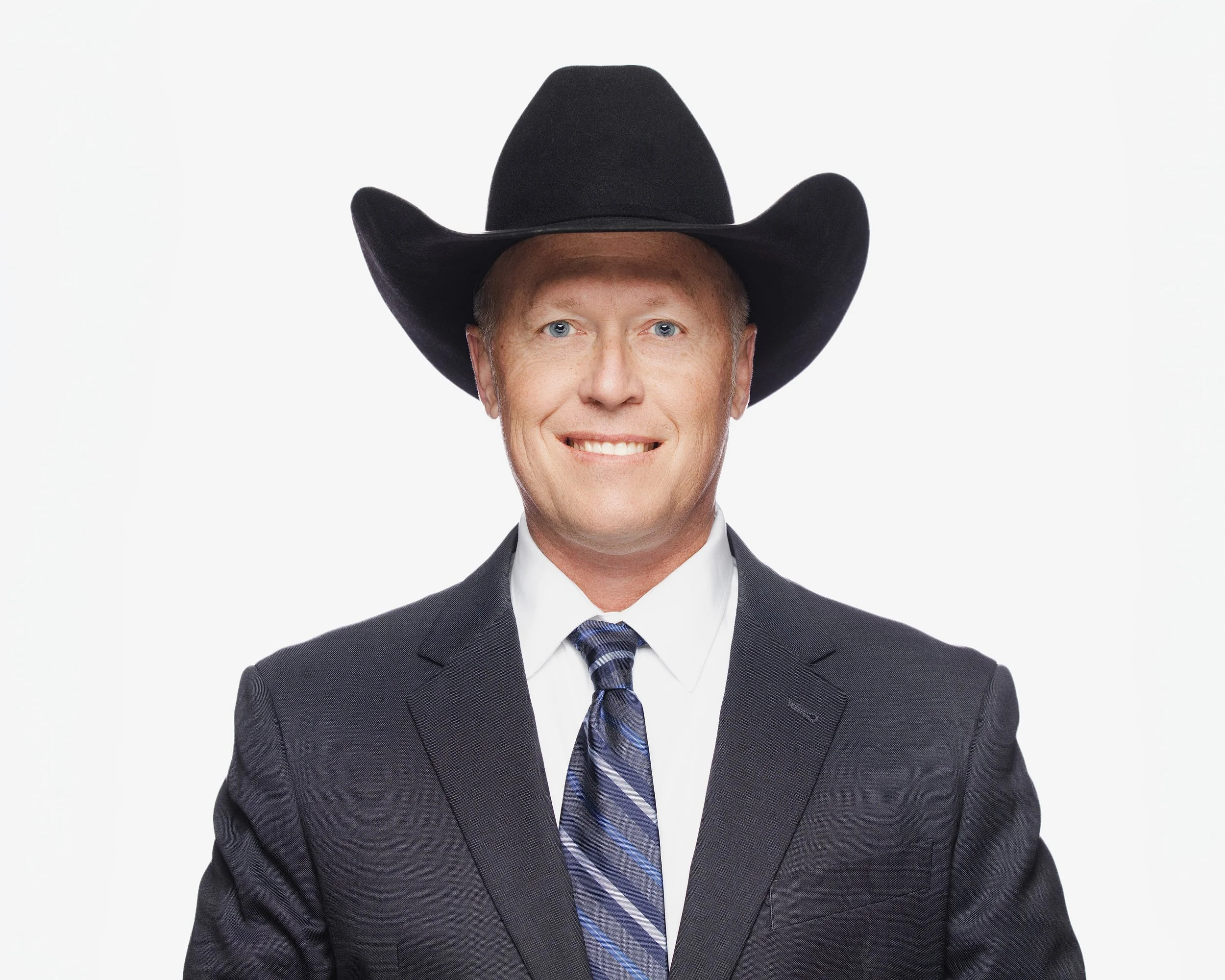 Headshot of a man in a suit and tie wearing a black cowboy hat against a plain white background.