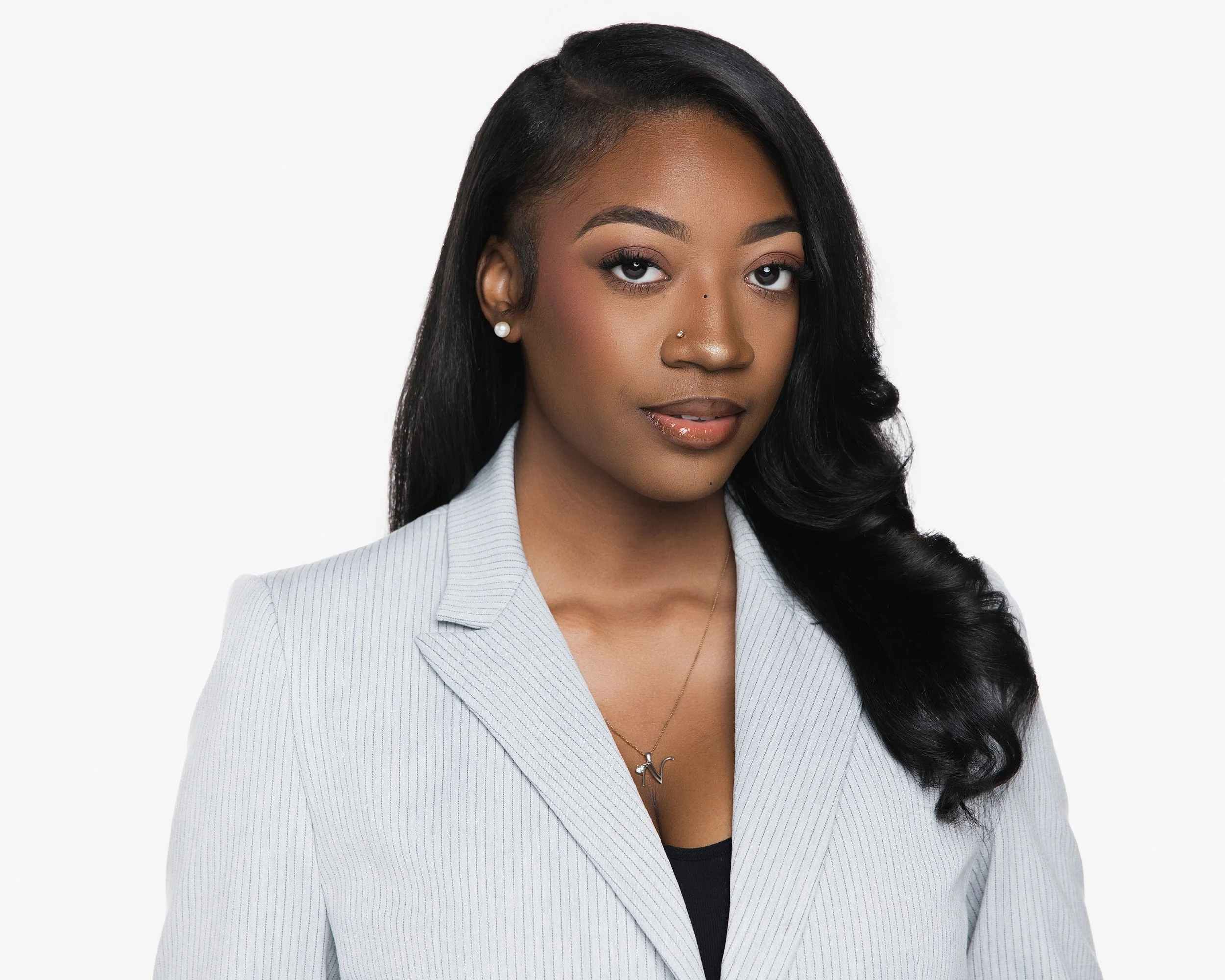 A woman with dark hair styled to one side, wearing pearl earrings and a silver necklace with a letter 'N'. She is dressed in a light gray pinstriped blazer over a black top, with makeup including eye makeup and lipstick, posing against a plain white background.