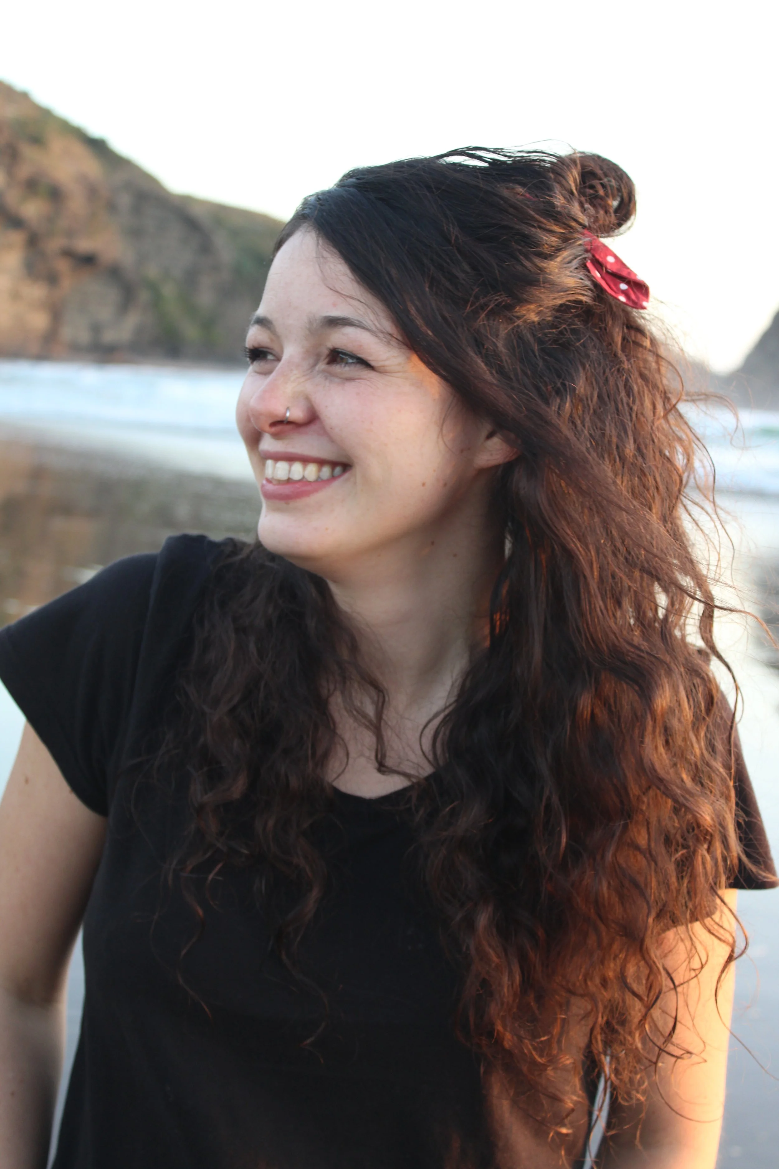 Young woman with long, curly brown hair and a nose piercing, smiling outdoors near a beach with cliffs in the background.
