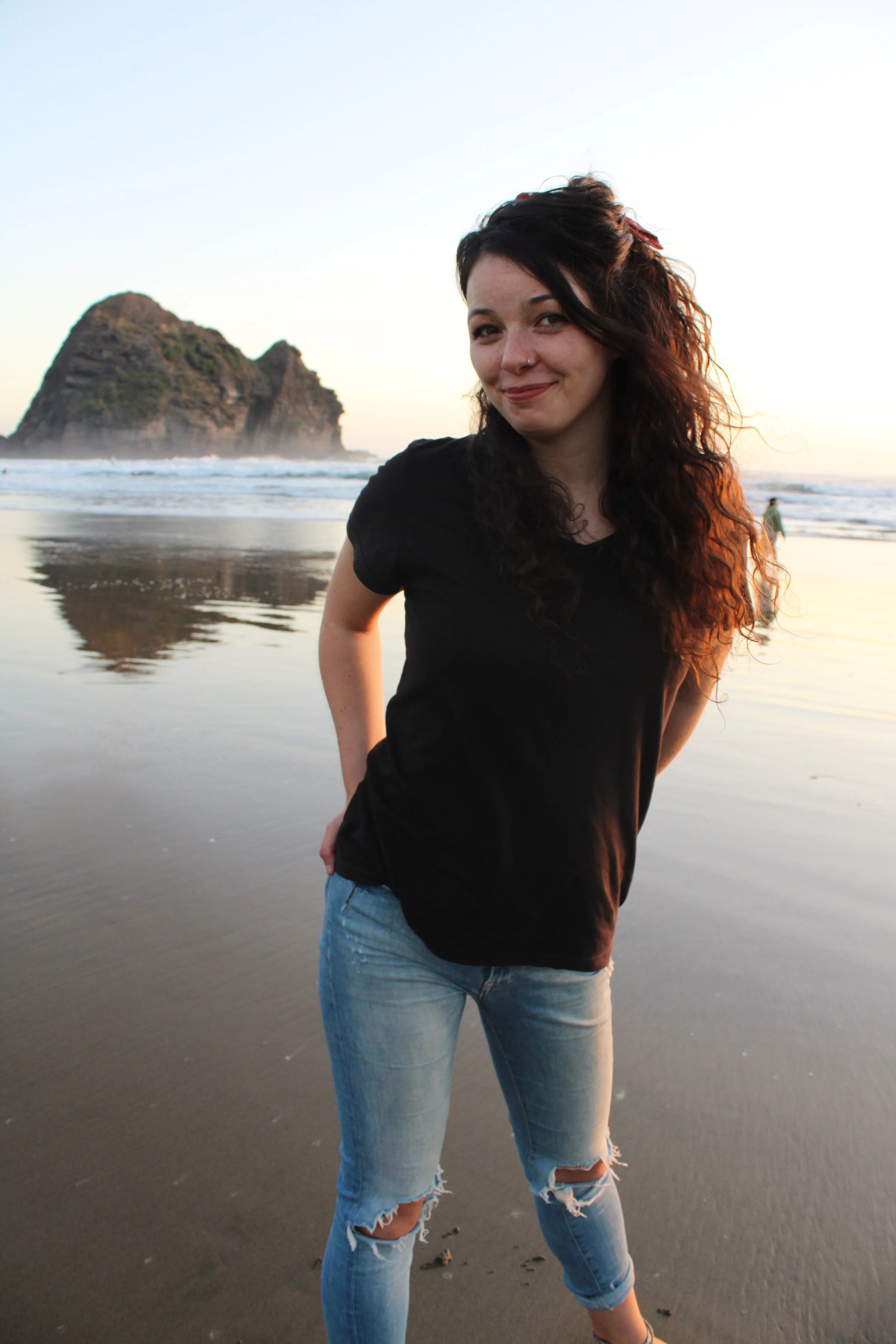 Young woman with curly hair and nose piercing at the beach during sunset, standing on wet sand with an ocean and rocky island in the background.