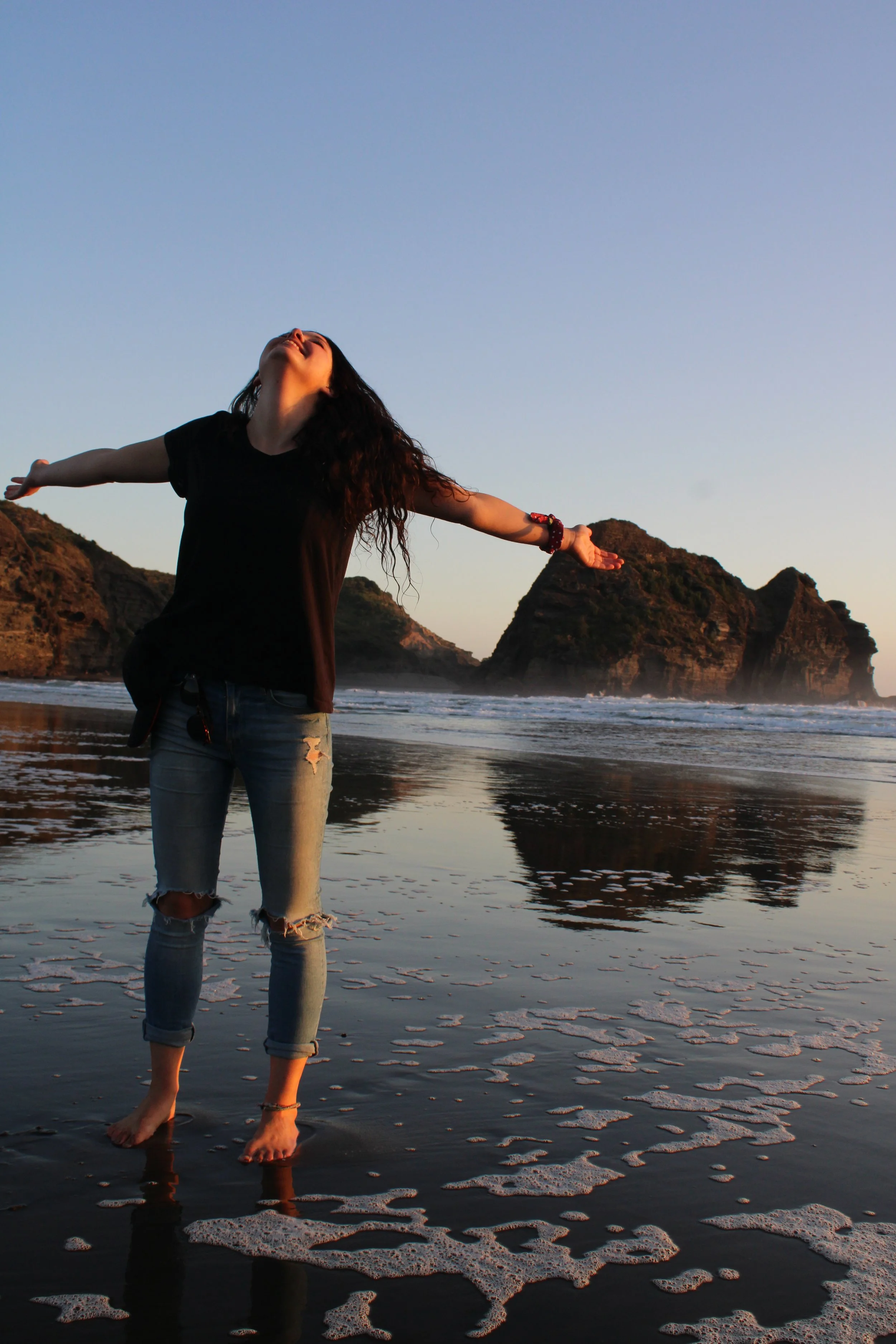 Woman standing in shallow ocean water with arms outstretched and head tilted back, waves and rocky islands in the background during sunset or sunrise.