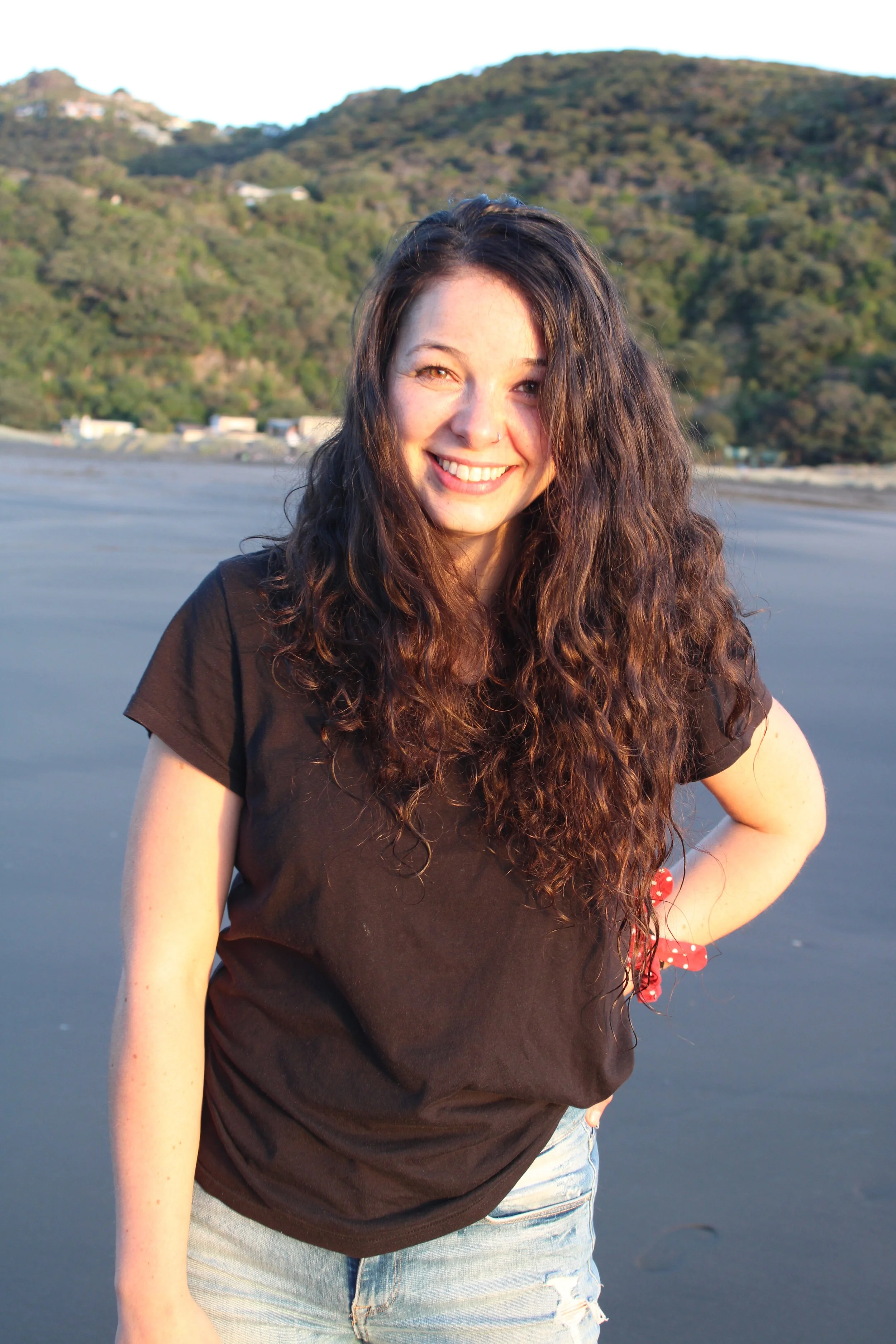 A young woman with long curly hair and a black t-shirt smiling on a beach with mountains in the background.