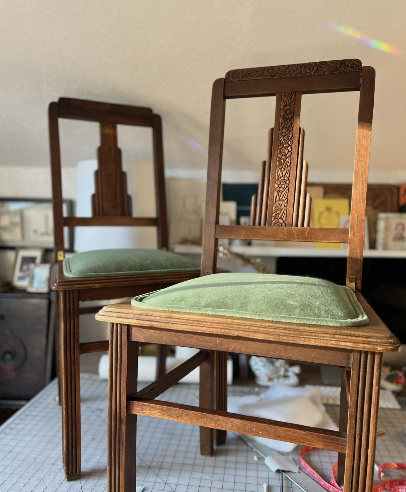 Two vintage wooden chairs with carved backrests and green cushioned seats, stacked one on top of the other in a room with various framed pictures and objects in the background.