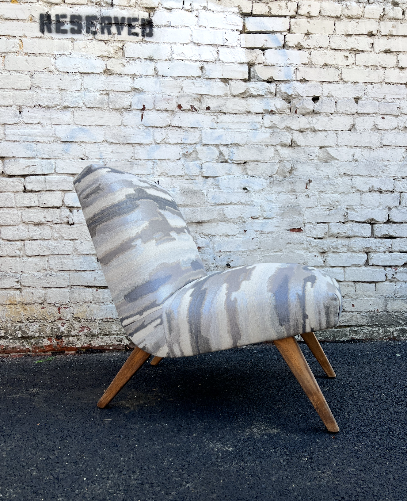An upholstered armchair with a curved back and wooden legs placed outdoors against a white brick wall with graffiti that reads 'RESERVE'.