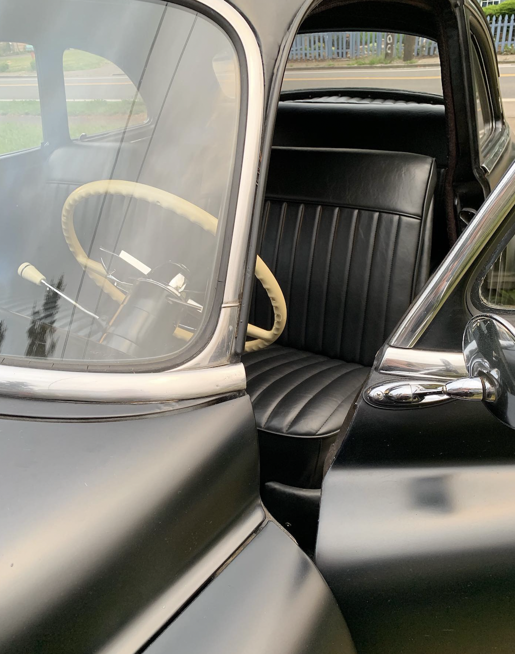 Close-up view of the front interior of a vintage black car, showing a black leather seat, a yellow steering wheel, and a windshield with a view of the road outside.