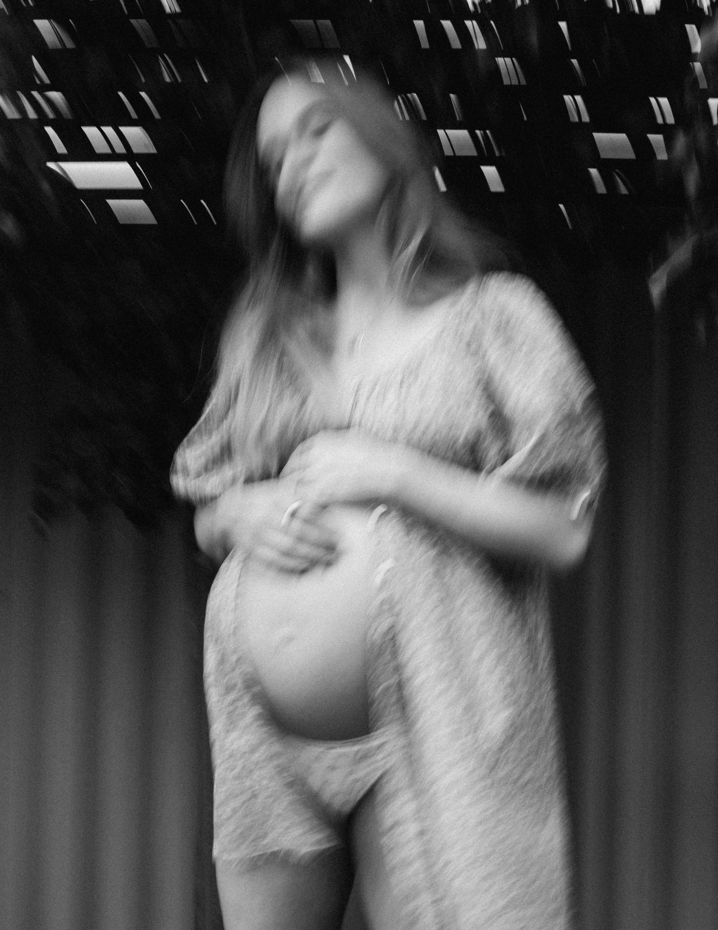 A pregnant woman standing against a backdrop of a cityscape at night, with blurred lights and windows. She is holding her belly with one hand and has her eyes closed, appearing peaceful.