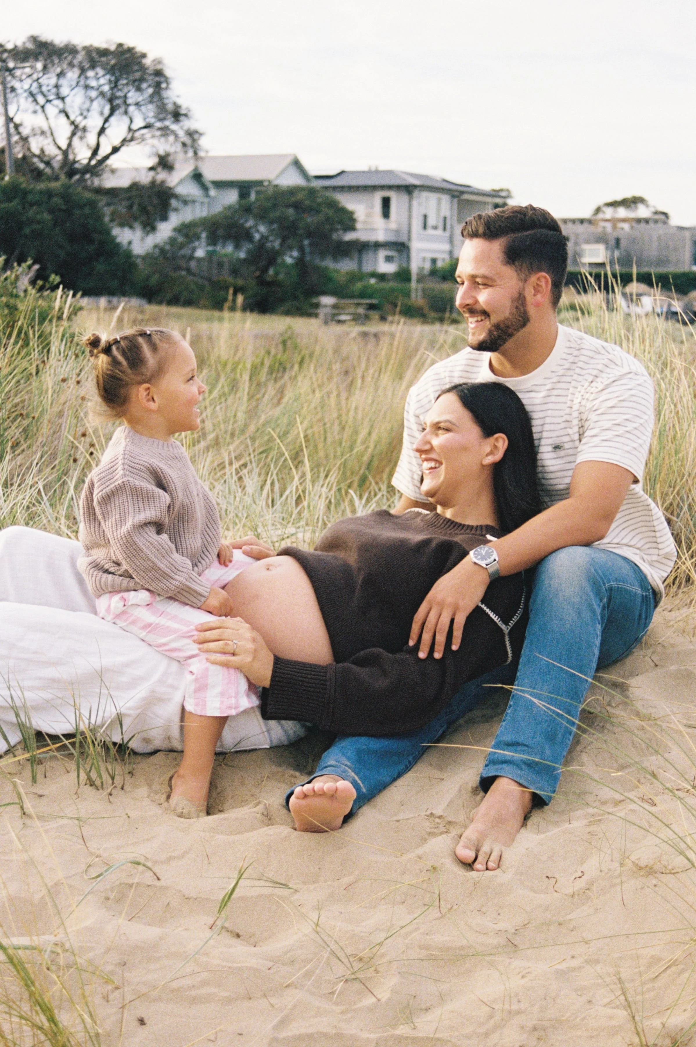 A family of four standing outdoors in a park during autumn, with yellow leaves on trees and fallen leaves on the grass, smiling and engaging with each other.