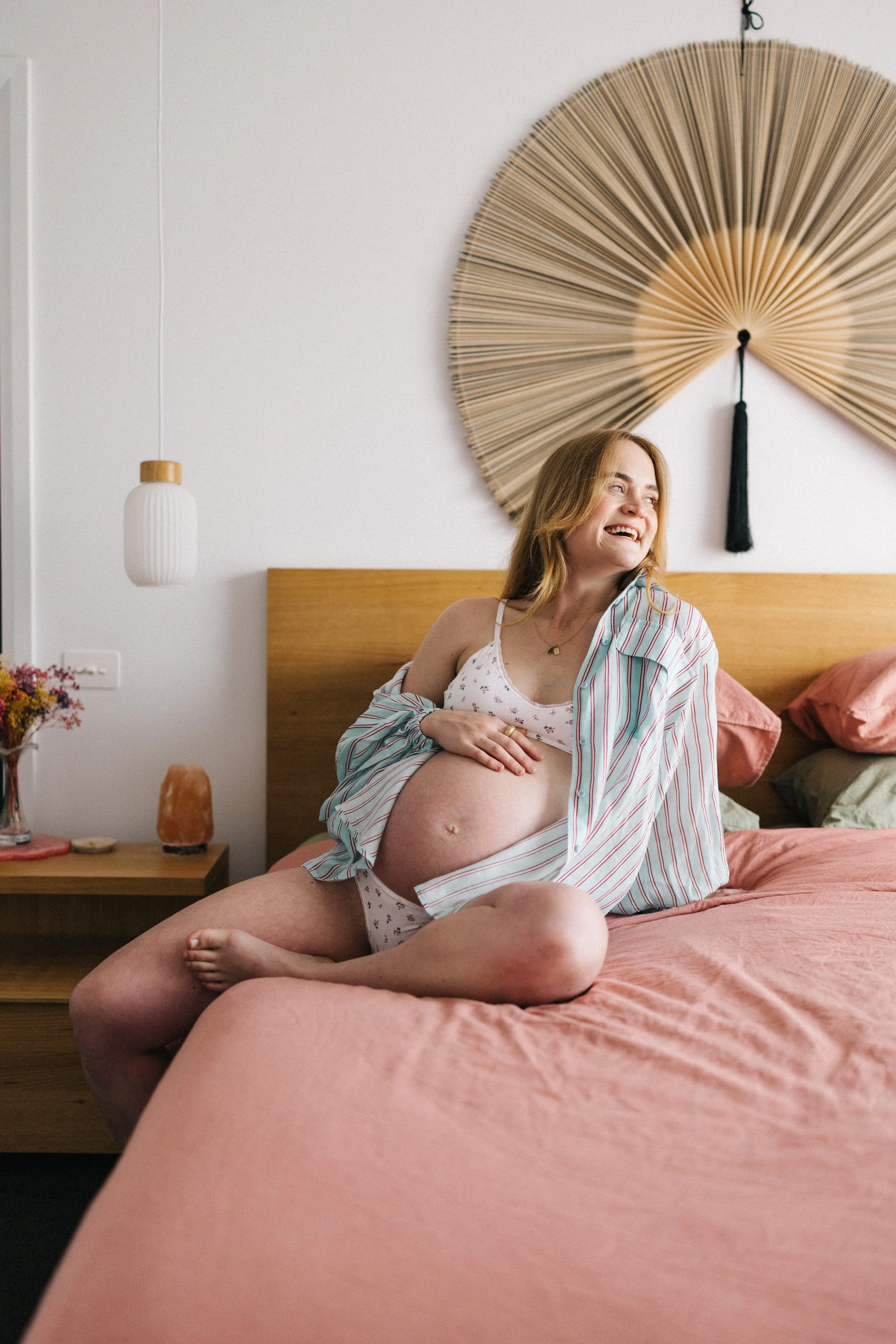 A pregnant woman sitting on a bed in a bedroom, smiling and looking to the side. She is wearing a striped shirt partially open, revealing her belly and lingerie with a floral pattern. The room has a wooden headboard, pink bedding, and decorative wall