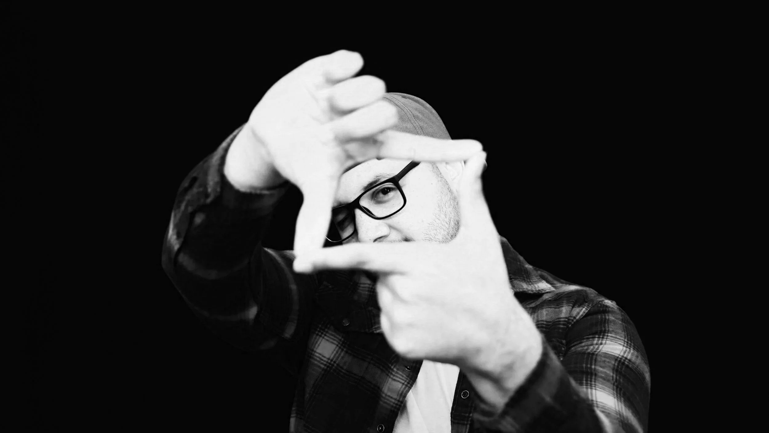 A man with glasses and a beard making a square frame with his hands, wearing a plaid shirt and a beanie, against a black background.