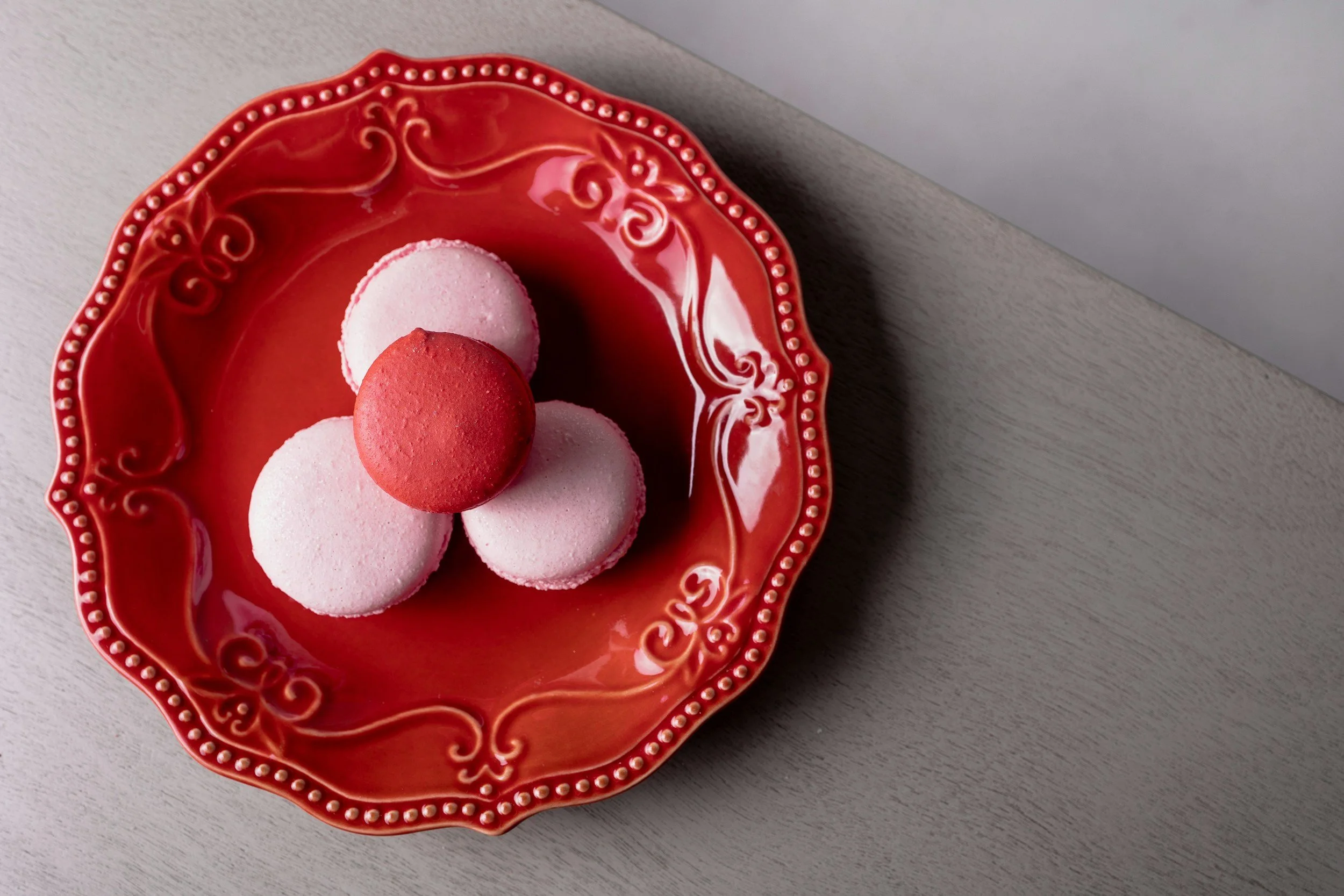 Red decorative plate with four round meringue cookies, one pink and one red on top, on a light-colored wooden surface.