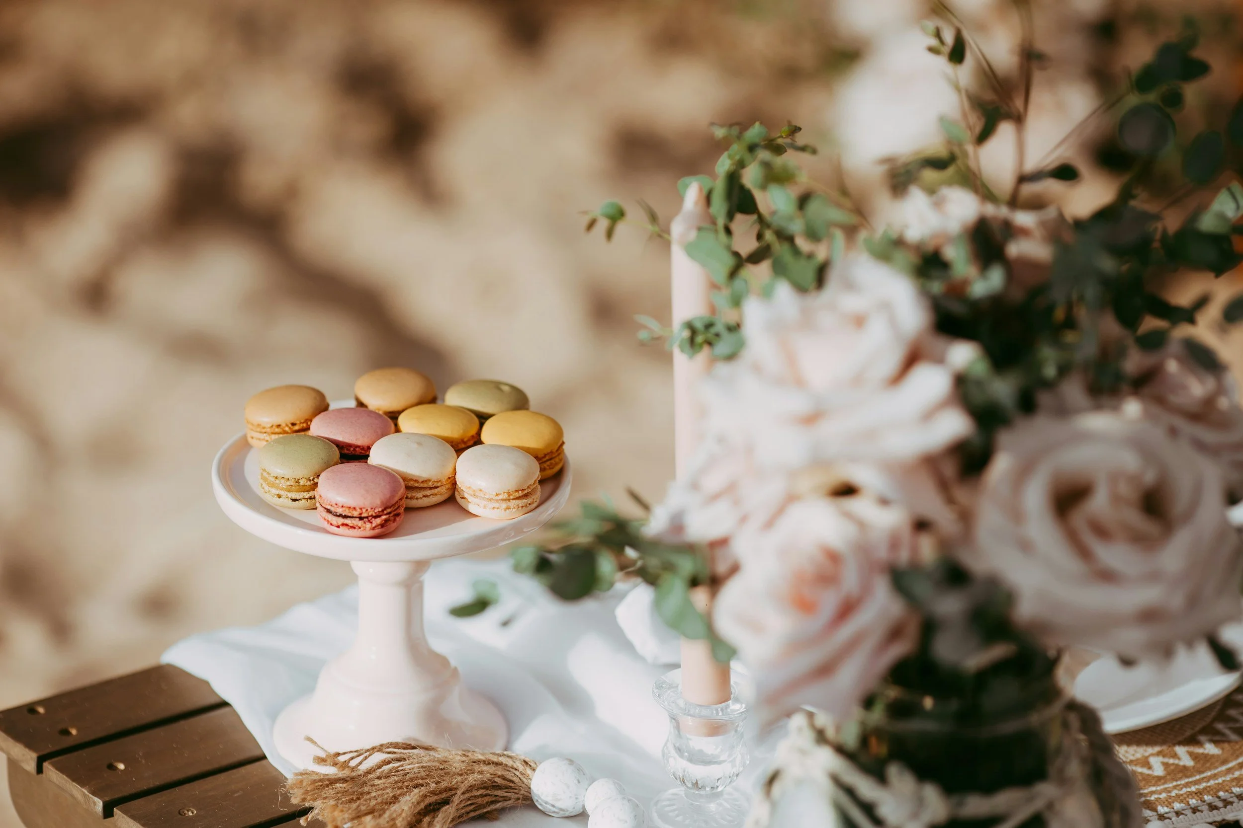 Colorful macarons on a white cake stand next to pink candles and floral decorations on a rustic table.