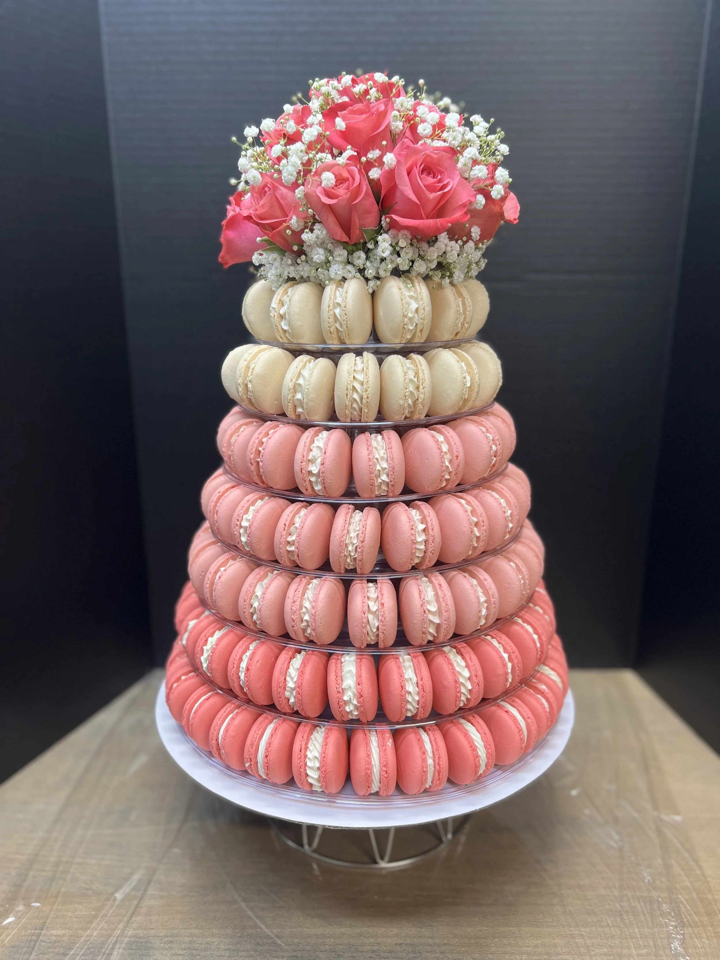 A multi-tiered cake stand decorated with pink and white macarons, topped with a bouquet of pink roses and small white flowers.
