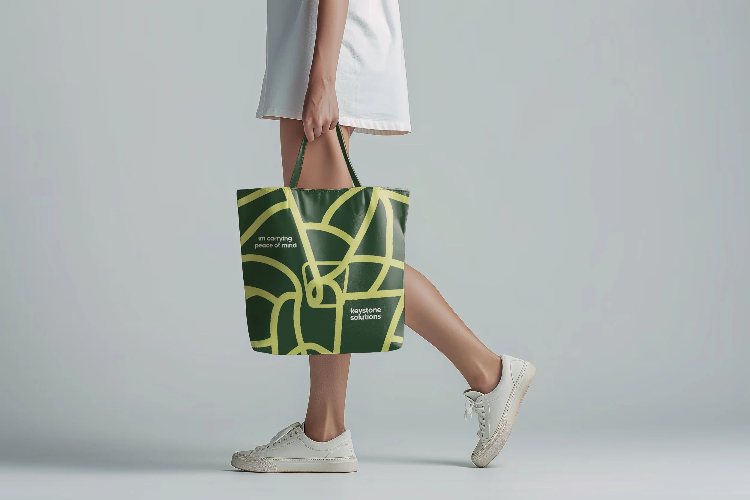Person holding a green reusable tote bag with yellow abstract lines and text, wearing a white dress and white sneakers, standing against a light gray background.