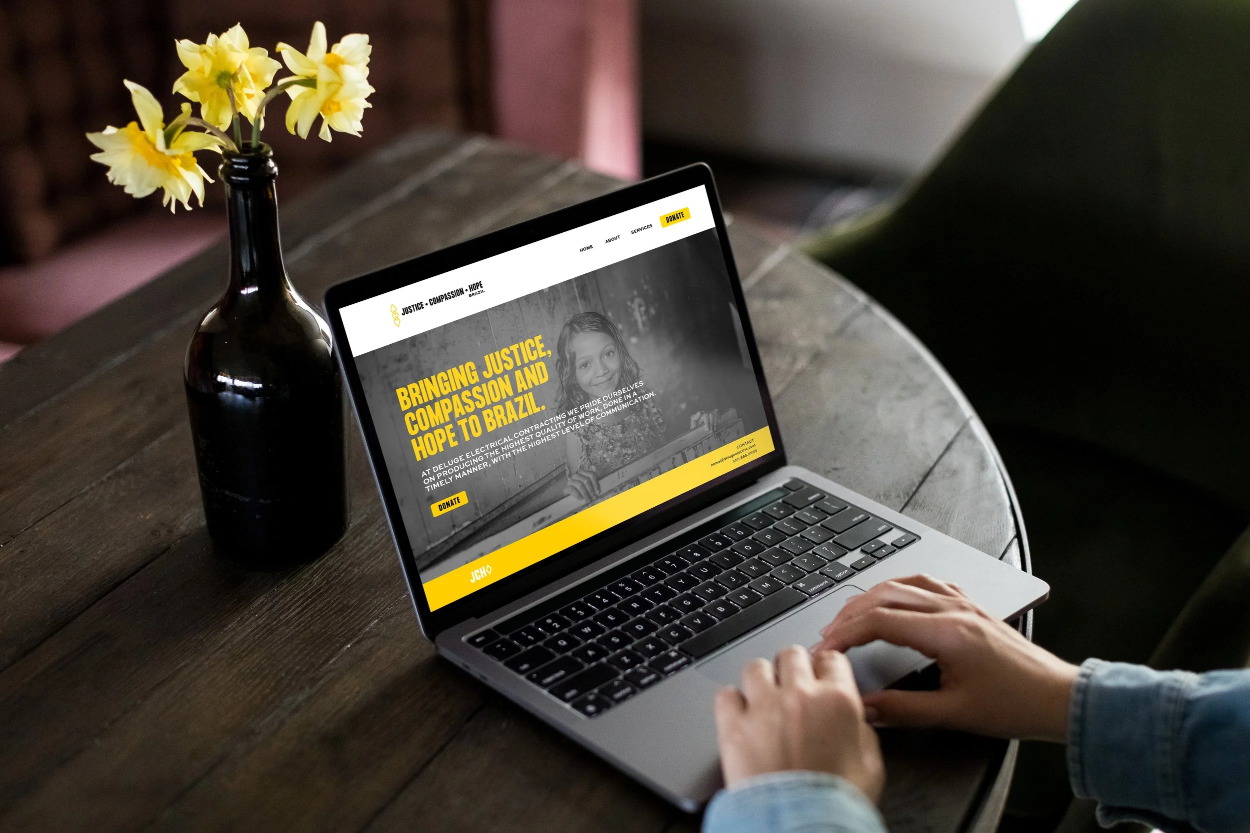 A person working on a laptop at a wooden table with a black vase of yellow flowers nearby. The laptop screen displays a website with a black and yellow color scheme and text promoting justice, compassion, and hope in Brazil.