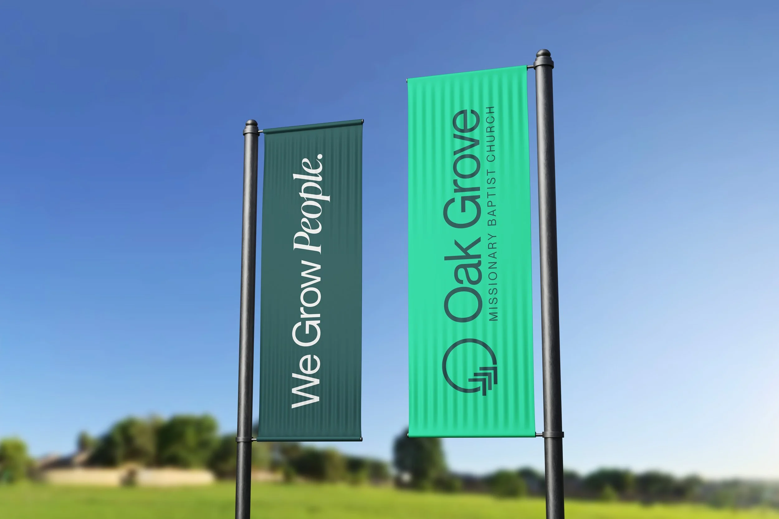 Two vertical banners on poles, one green and one teal, displaying promotional text for a church event, set against a blue sky with blurred background of trees and grass.