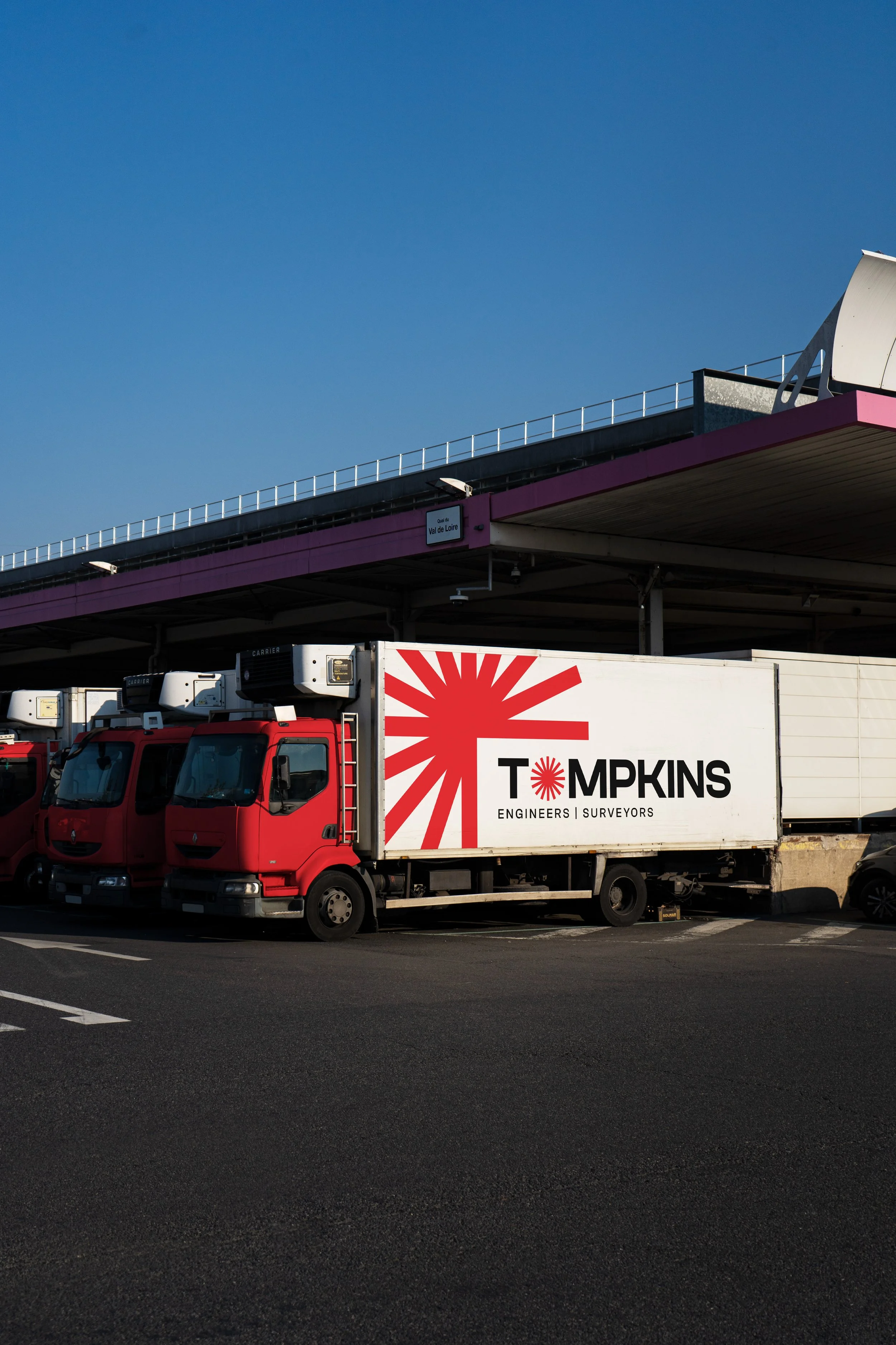 A delivery truck parked in a lot with the logo 'Tompkins' and the words 'Engineers | Surveyors' on the side, under an overpass with a purple edge, against a clear blue sky.