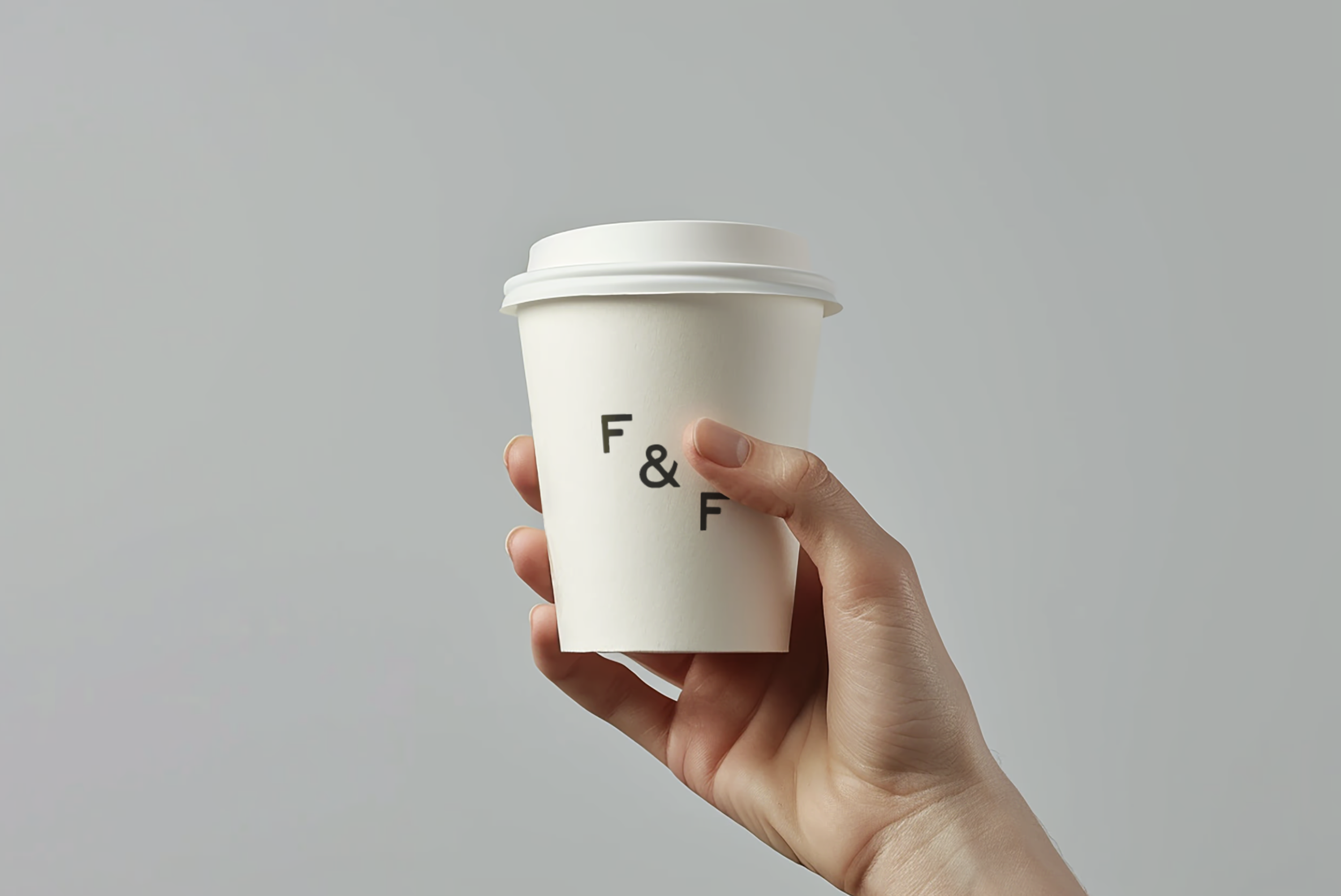 Hand holding a white disposable coffee cup with a black text logo against a plain light gray background.