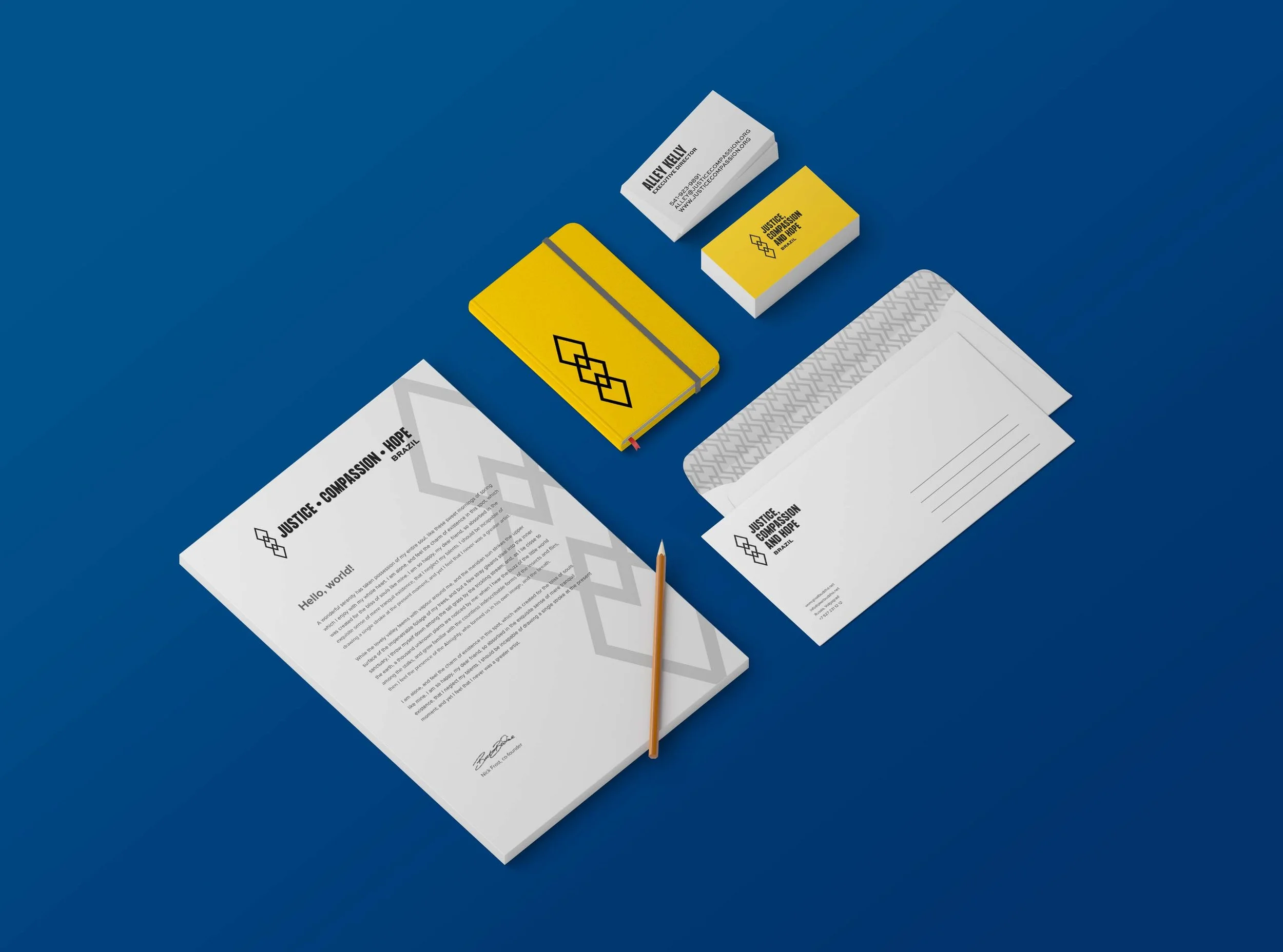 A collection of stationery items on a blue surface, including a yellow notebook, a pencil, a white letter, and business cards with the logo and text for Justice, Compassion, and Hope in Brazil.