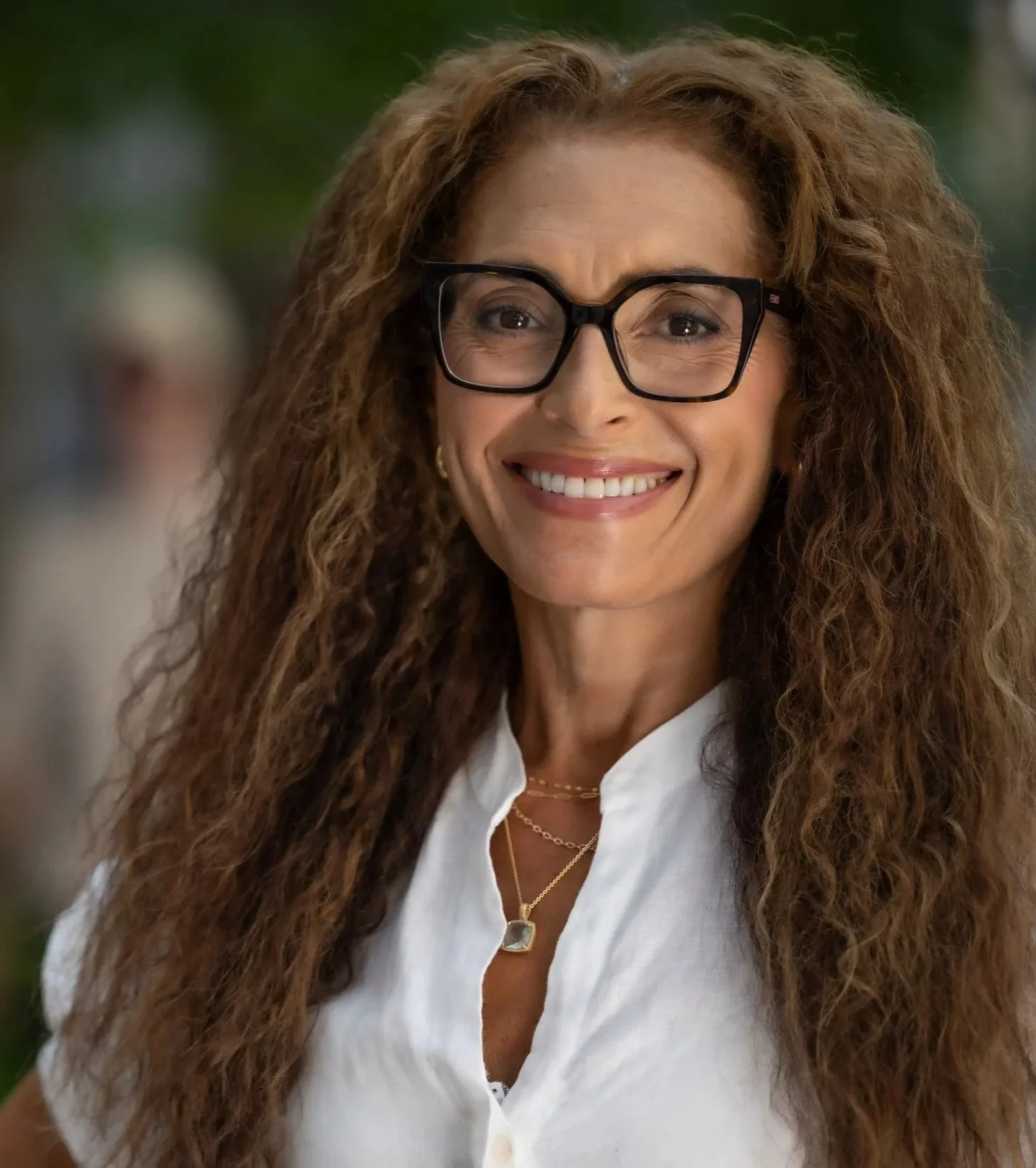 Close-up of a smiling woman with long curly brown hair wearing black glasses, a white shirt, and gold jewelry, outdoors with a blurred green background.