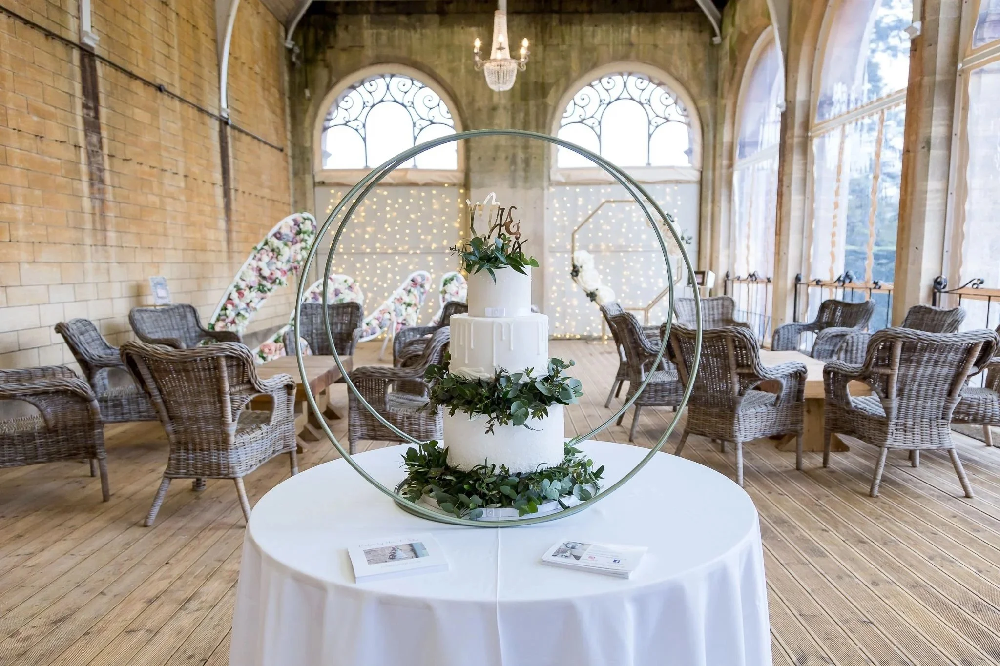 A three-tier iced wedding cake, decorated with greenery with metallic topper and surrounded by a double hoop. It is displayed on a round table with white tablecloth in the Orangery at Grittleton House
