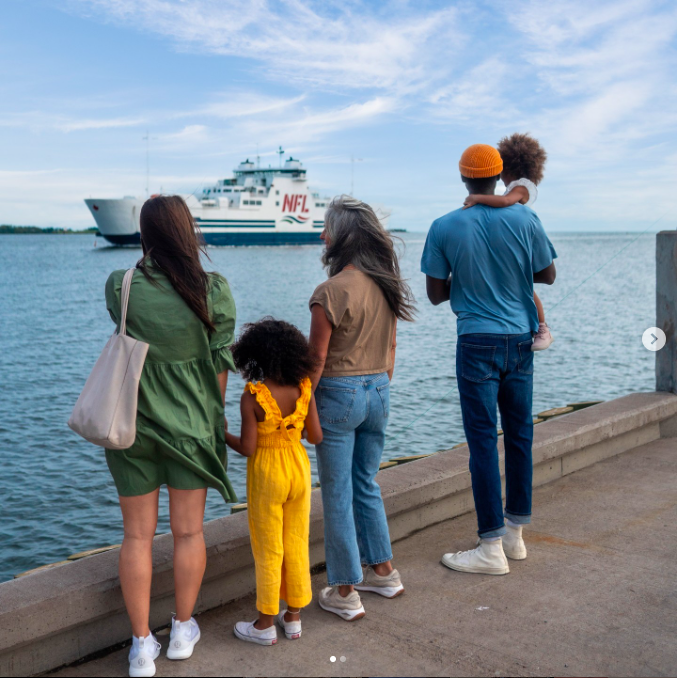 A group of people, including two adults and two children, stand on a pier watching a large ferry with "NFL" written on it pass by in the water. The sky is partly cloudy and the atmosphere appears calm and scenic.