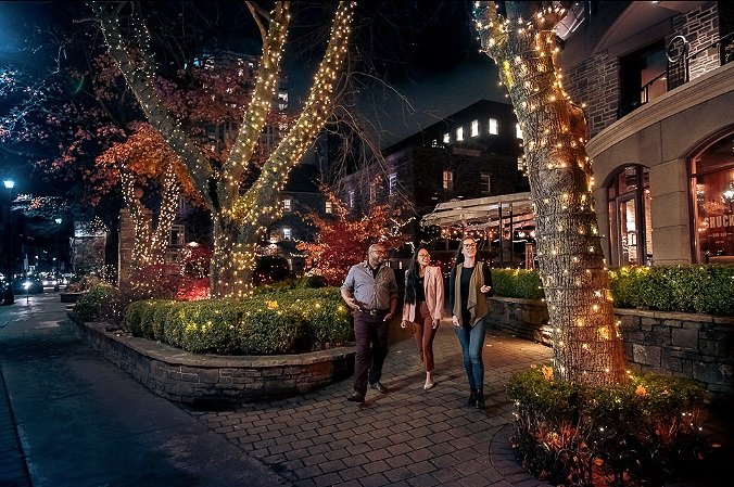 Three people walking on a brick sidewalk at night with trees adorned with string lights and illuminated buildings in the background.