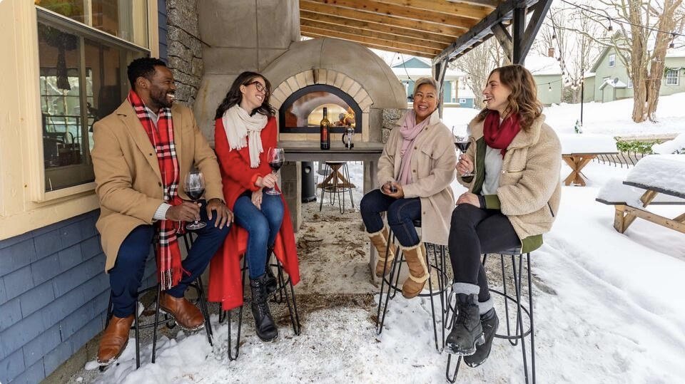 Four people sitting on stools outside next to a wood-fired pizza oven, with snow on the ground, holding wine glasses and smiling.