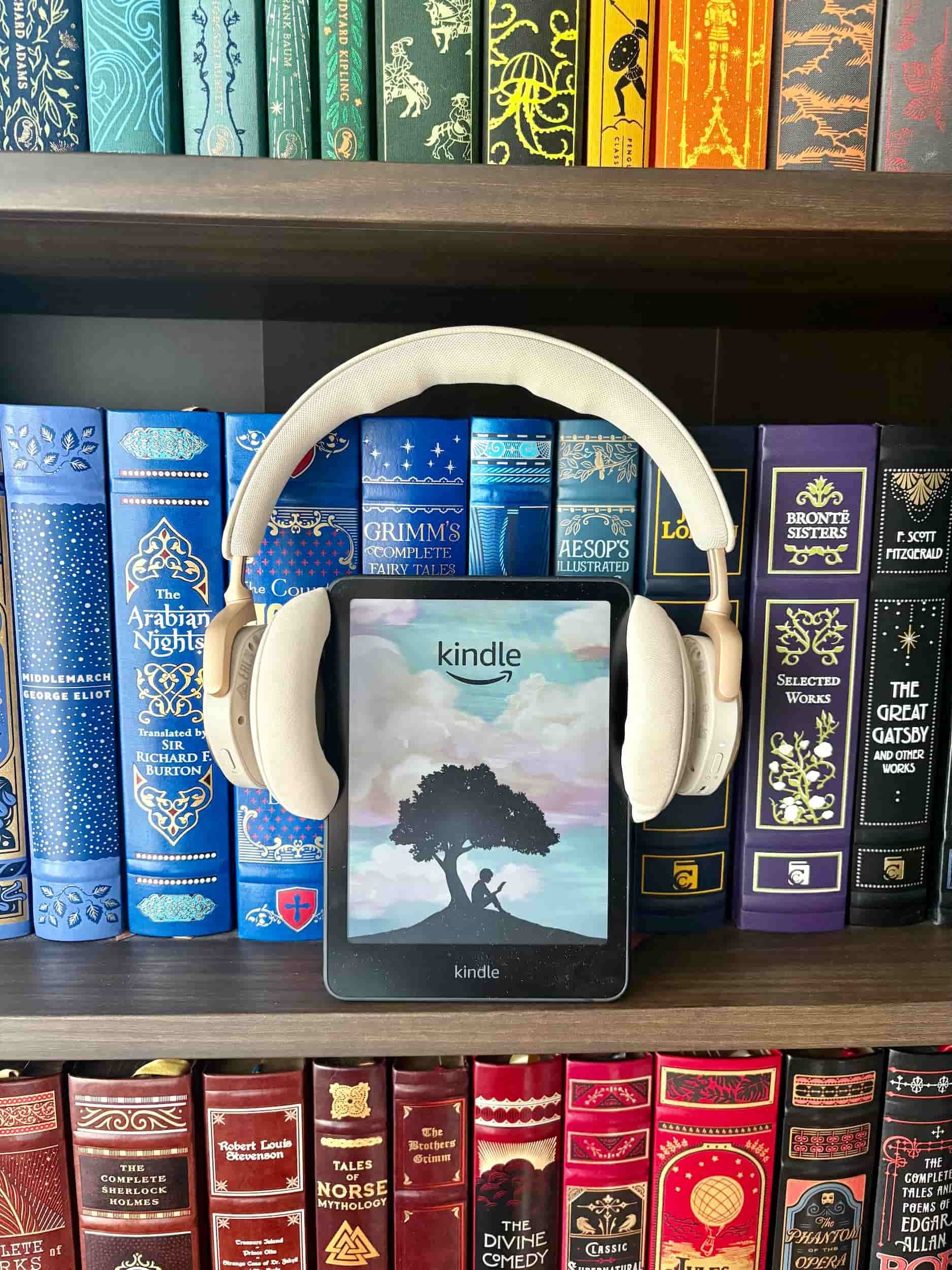 A Kindle with Headphones on, standing atop a bookshelf with multi coloured books, signifying how switching to audio books can help readers get out of a reading slump.