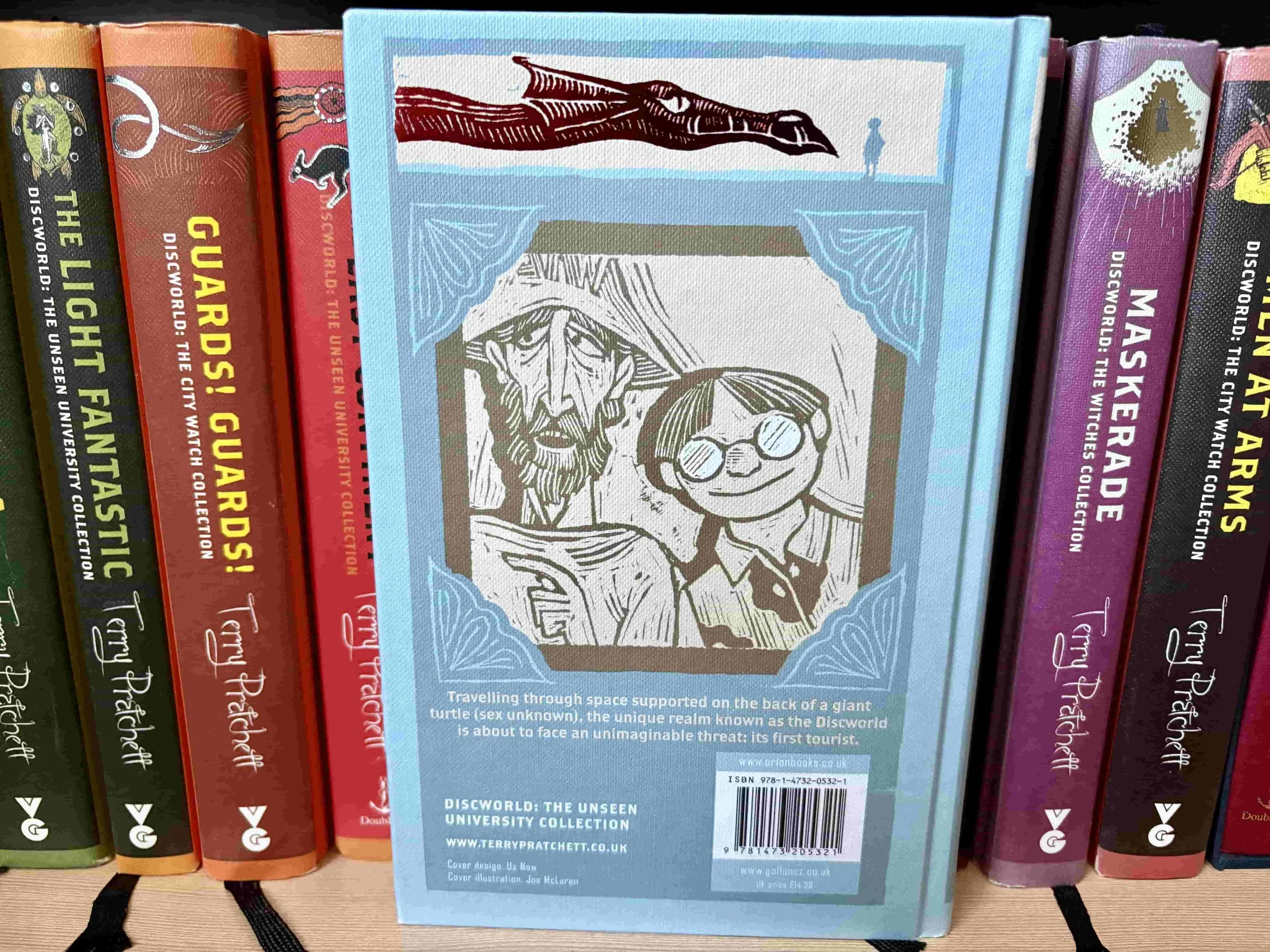 The back cover of the book The Colour of Magic by Terry Pratchett next to other books from the Discworld series, photo taken from the personal library of His and Hers Book Club