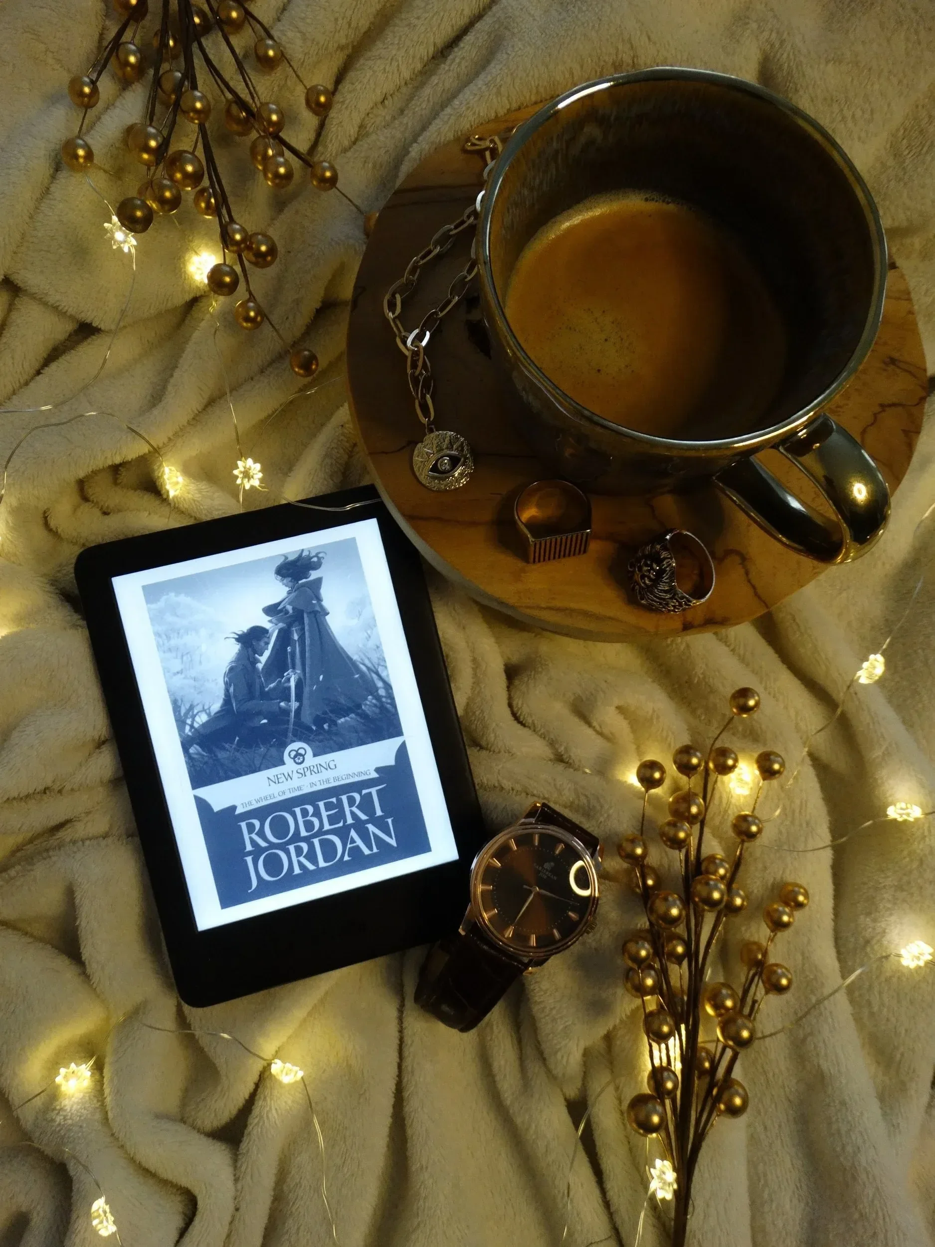 Robert Jordan's The Wheel of Time books on a Kindle, sitting atop a being blanket, next to a classic watch and decoration lights, with a cup of coffee above it.