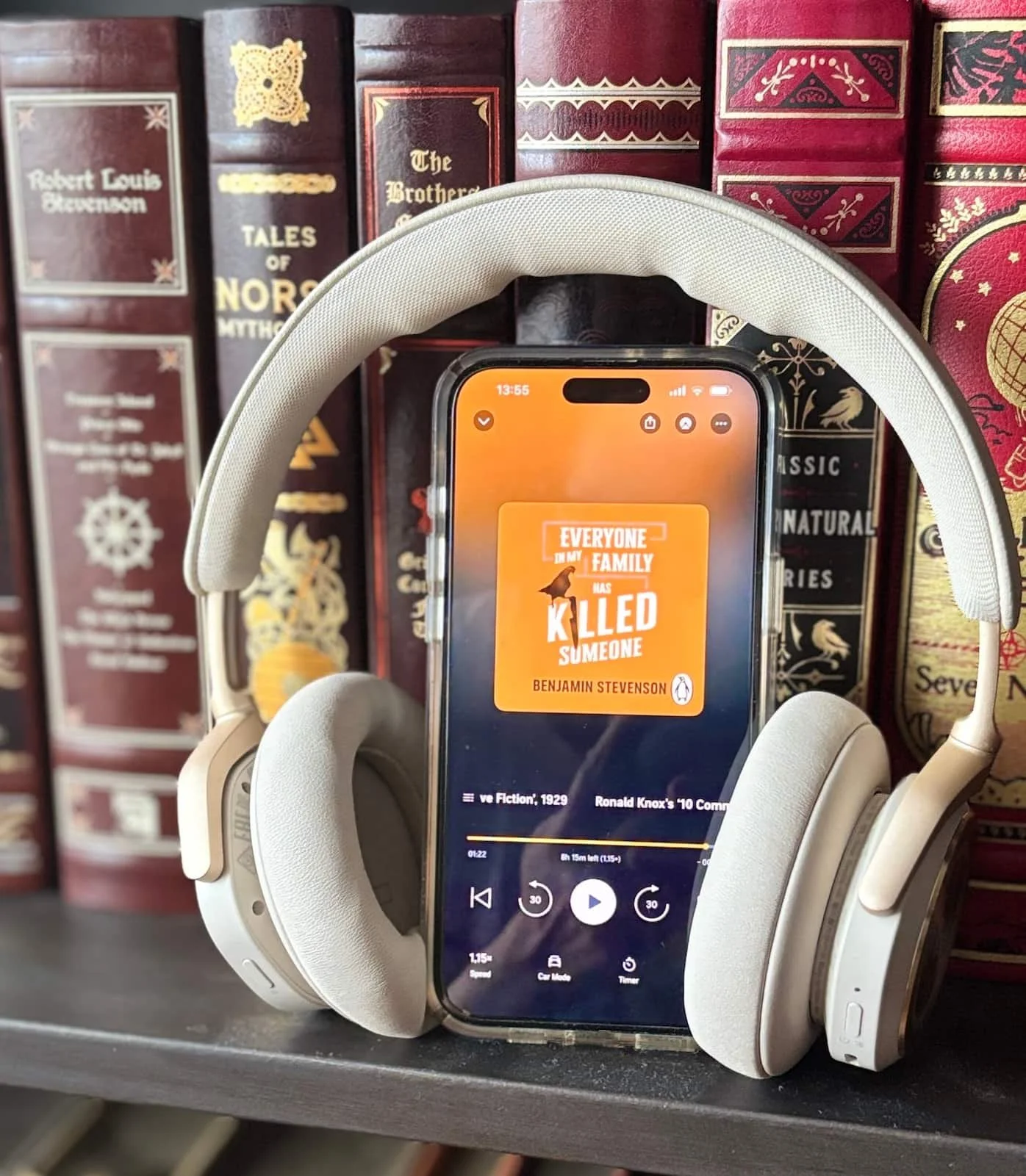 The audio book Everyone in my family has killed someone by Benjamin Stevenson, loaded on an iphone, which is sitting on a bookshelf, with headphones attached to it.