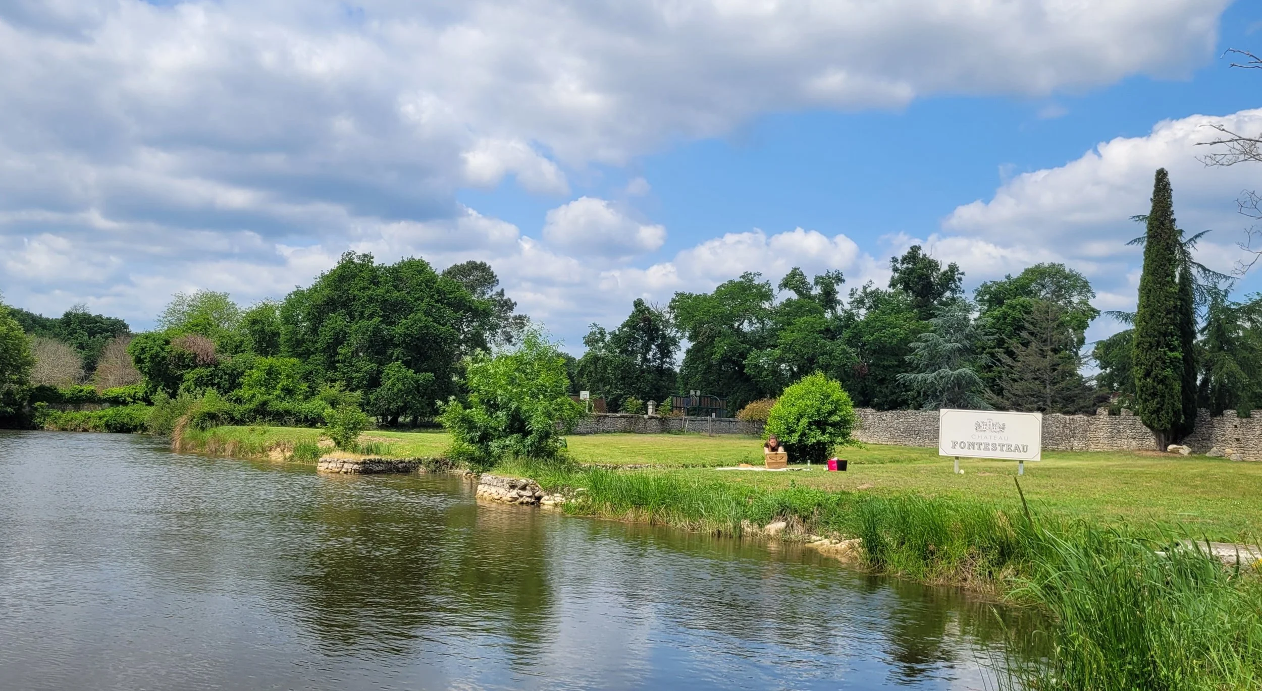 Paysage avec une rivière, des arbres verts, un ciel nuageux, un panneau indiquant "Fontesteau", et un mur de pierre.