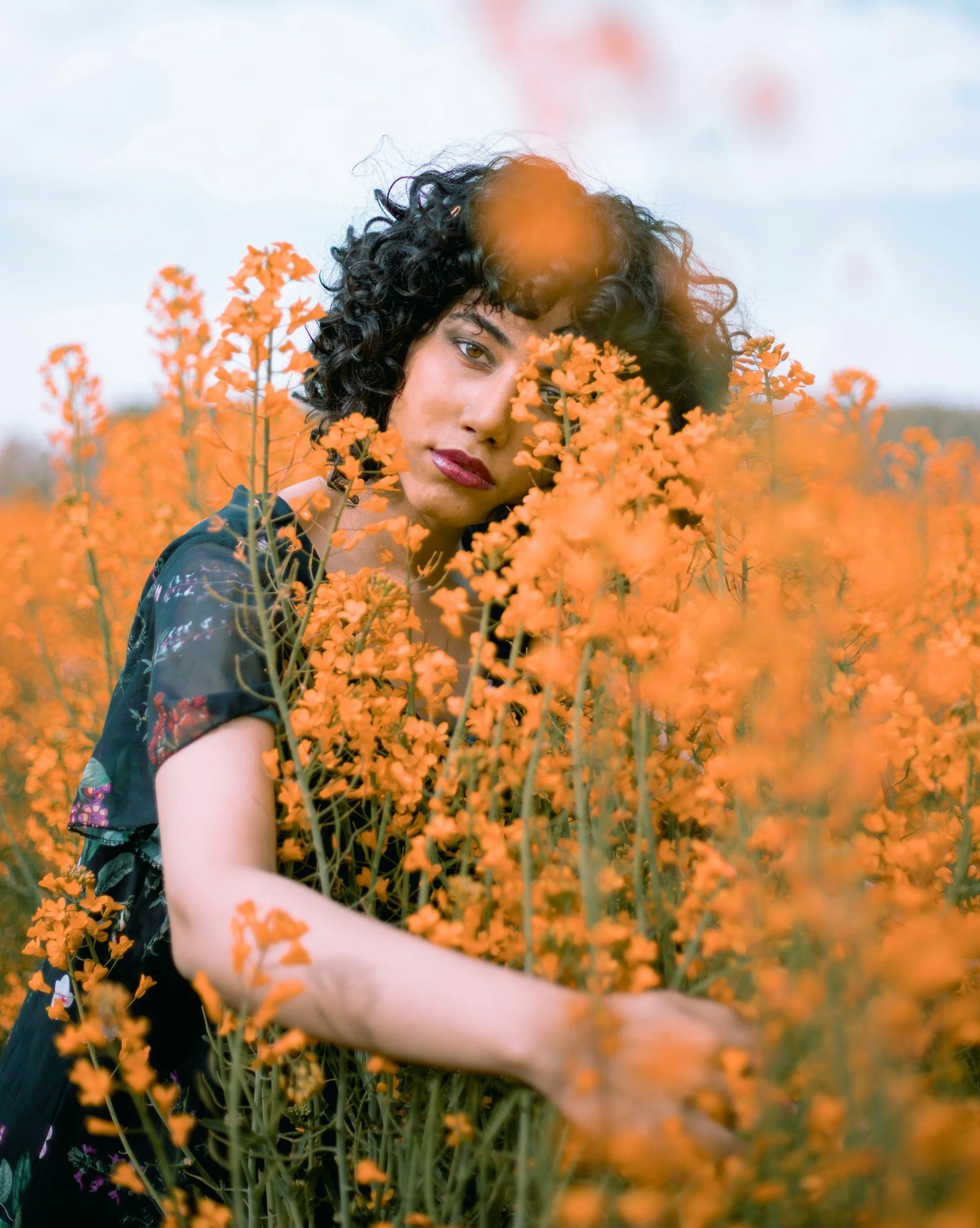 Woman with dark curly short hair and red lipstick sitting in a field of yellowish orange flowers where only half of her face is visible