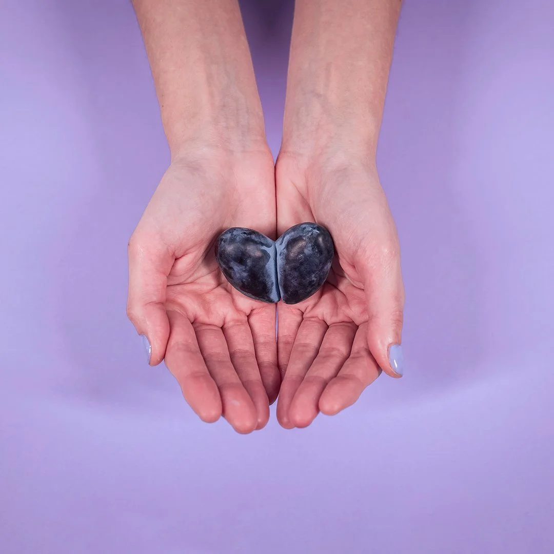 Hands holding a heart-shaped plum split in half against a lavender background, reflecting Plumline's playful visual identity.