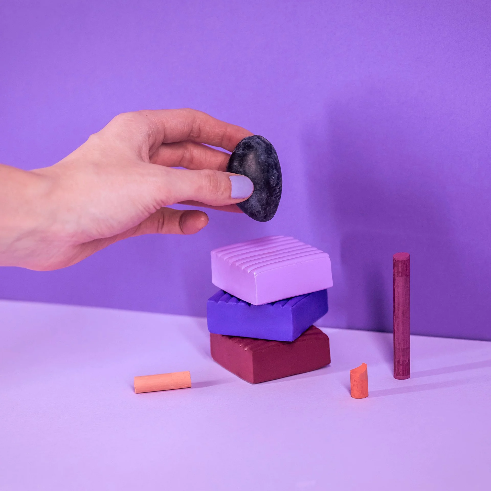 A hand positioning a plum above stacked purple and burgundy geometric objects against a lavender background.