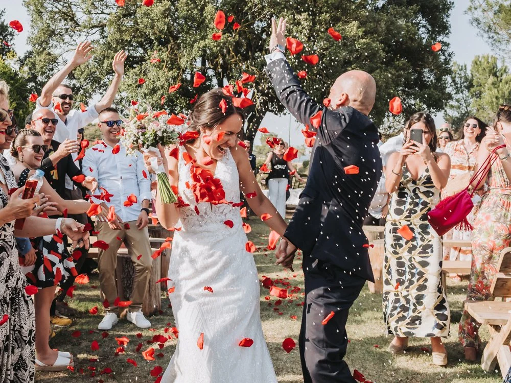 A bride and groom celebrate their wedding with guests throwing rose petals at them outdoors on a sunny day.
