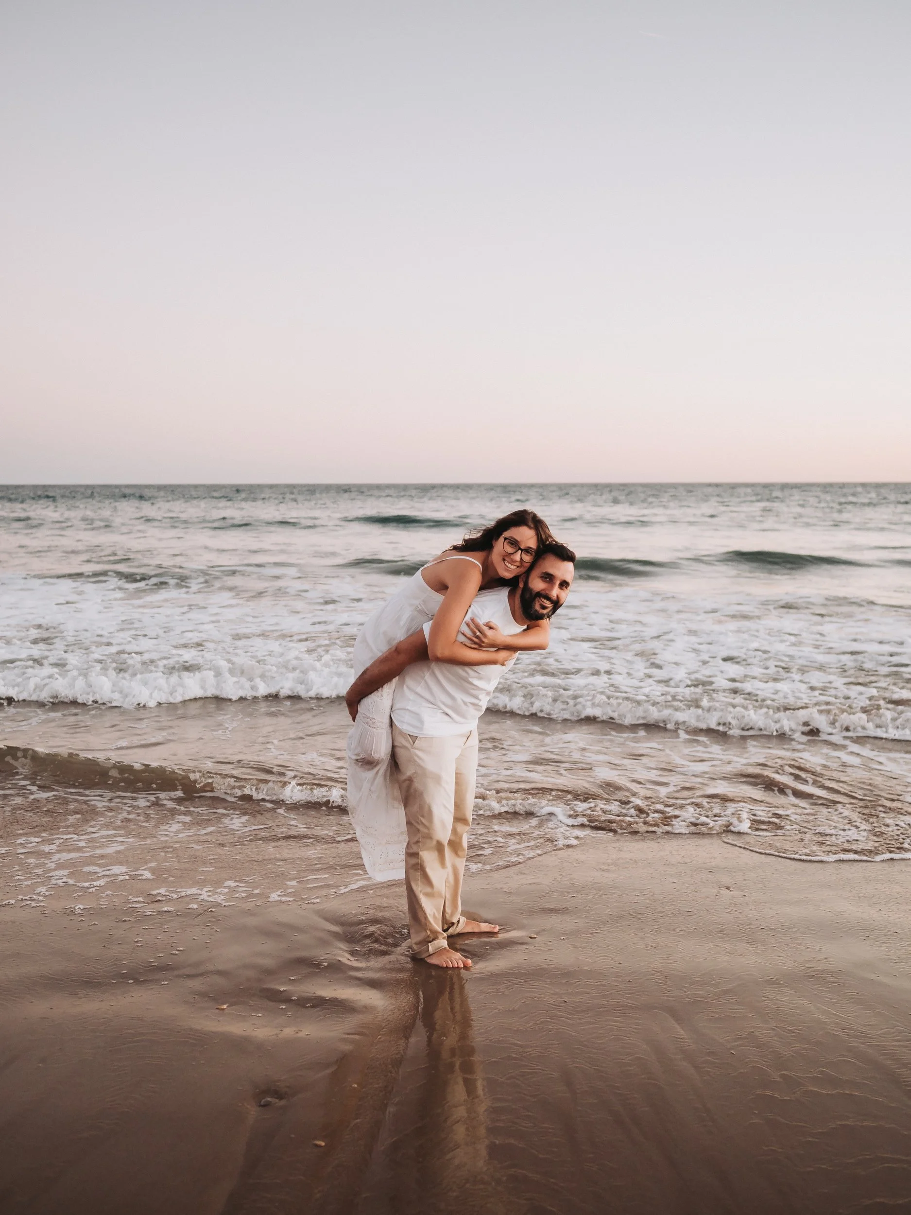 A happy couple at the beach, with the woman on the man's back, both smiling, near the waves and sandy shore at sunset.