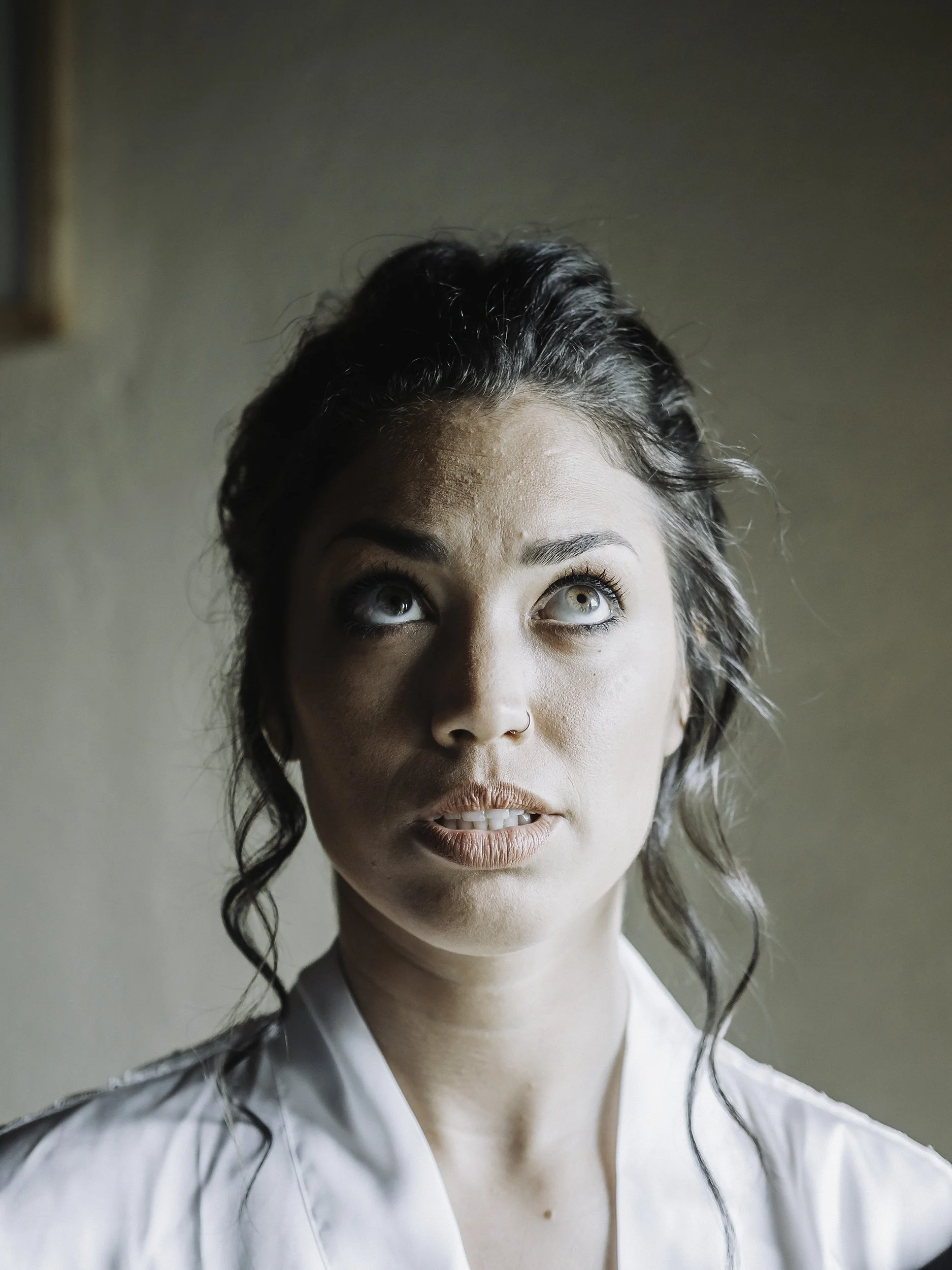 A woman with dark curly hair styled in an updo, light skin, blue eyes, wearing a nose ring, and a white shirt, looking upward with a neutral expression.
