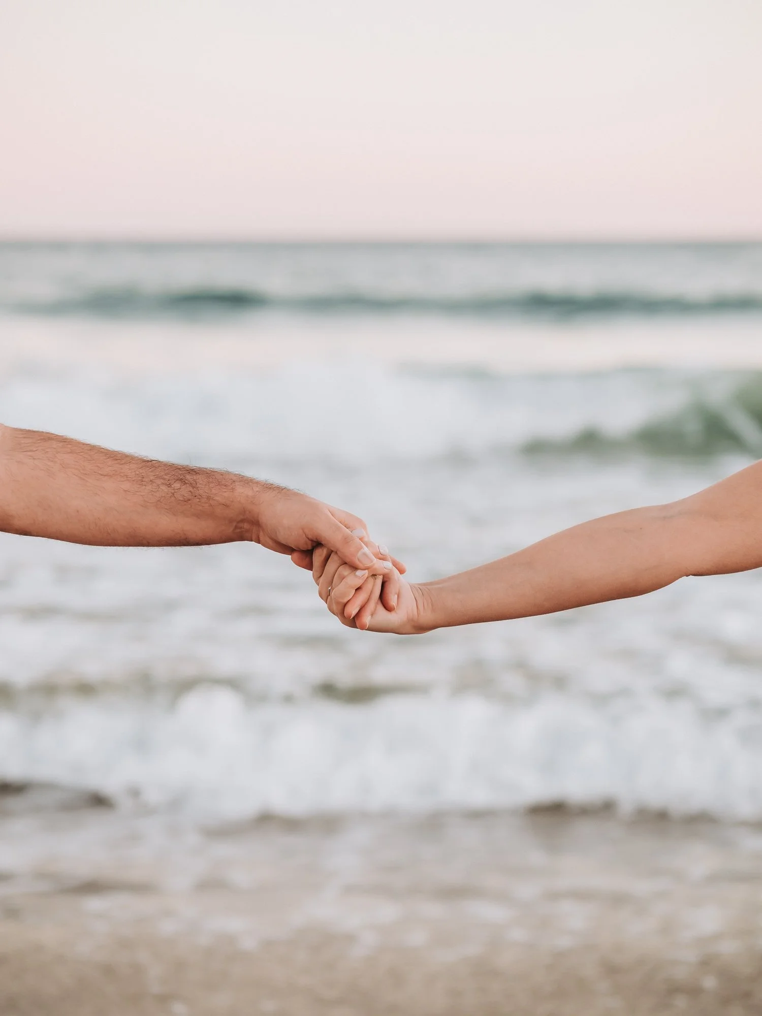 Hands holding each other on the beach, ocean waves in the background, during daylight.
