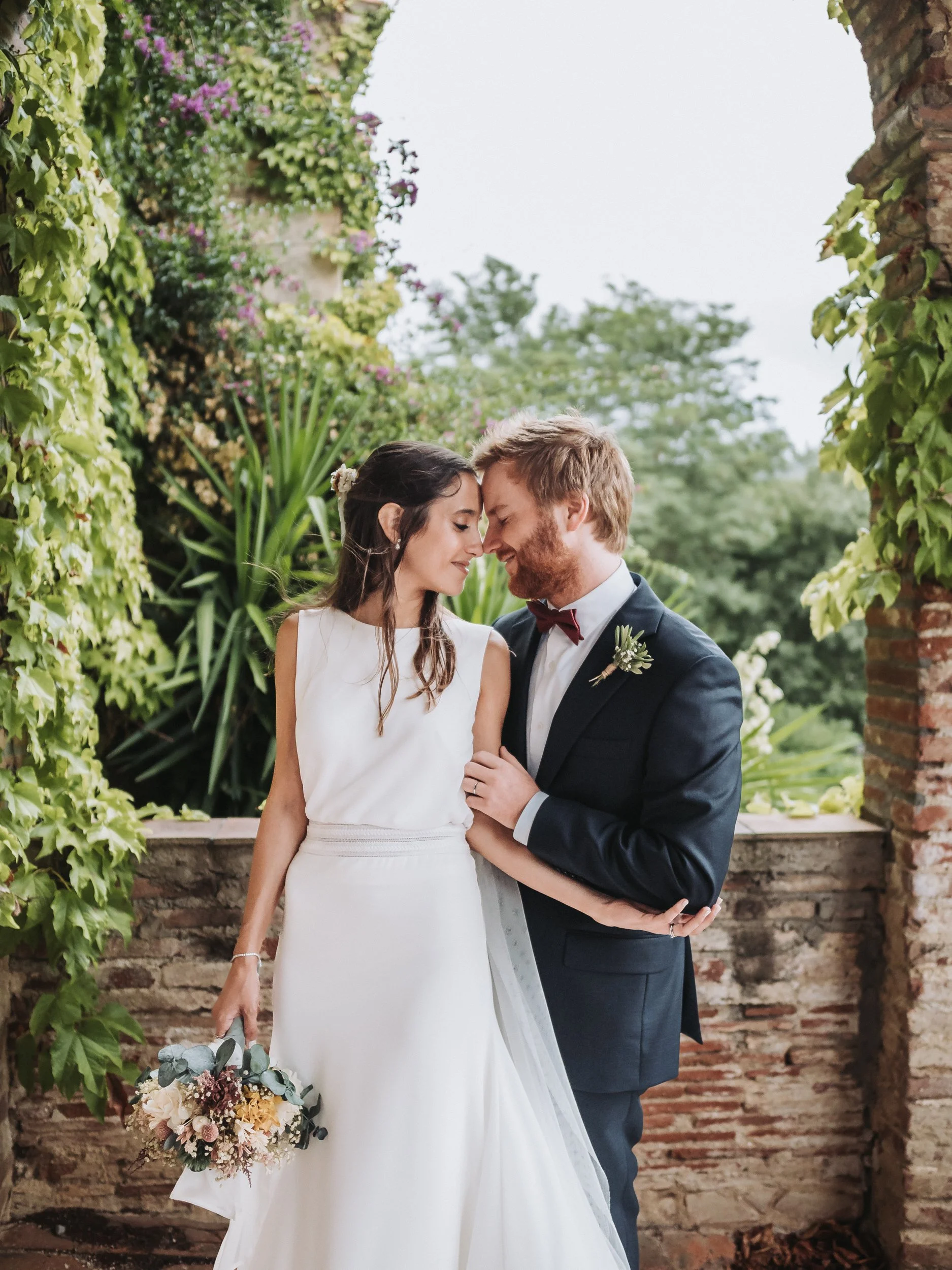 A bride and groom standing close with foreheads touching, smiling gently, in an outdoor garden setting with lush green plants and a brick wall.