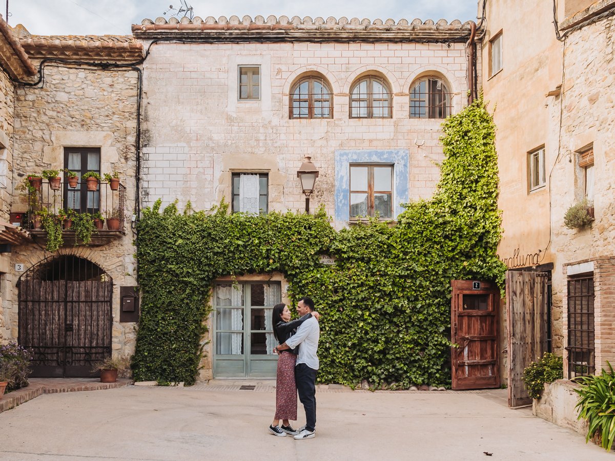 A couple is dancing and embracing in a courtyard of an old European-style stone building with green ivy, potted plants, and arched windows.