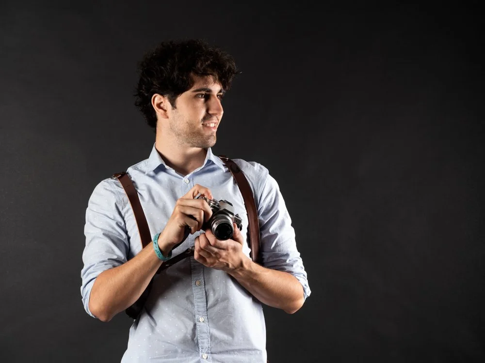 Young man with dark, curly hair, wearing a light blue shirt with rolled-up sleeves and brown suspenders, holding a camera, standing against a dark background.