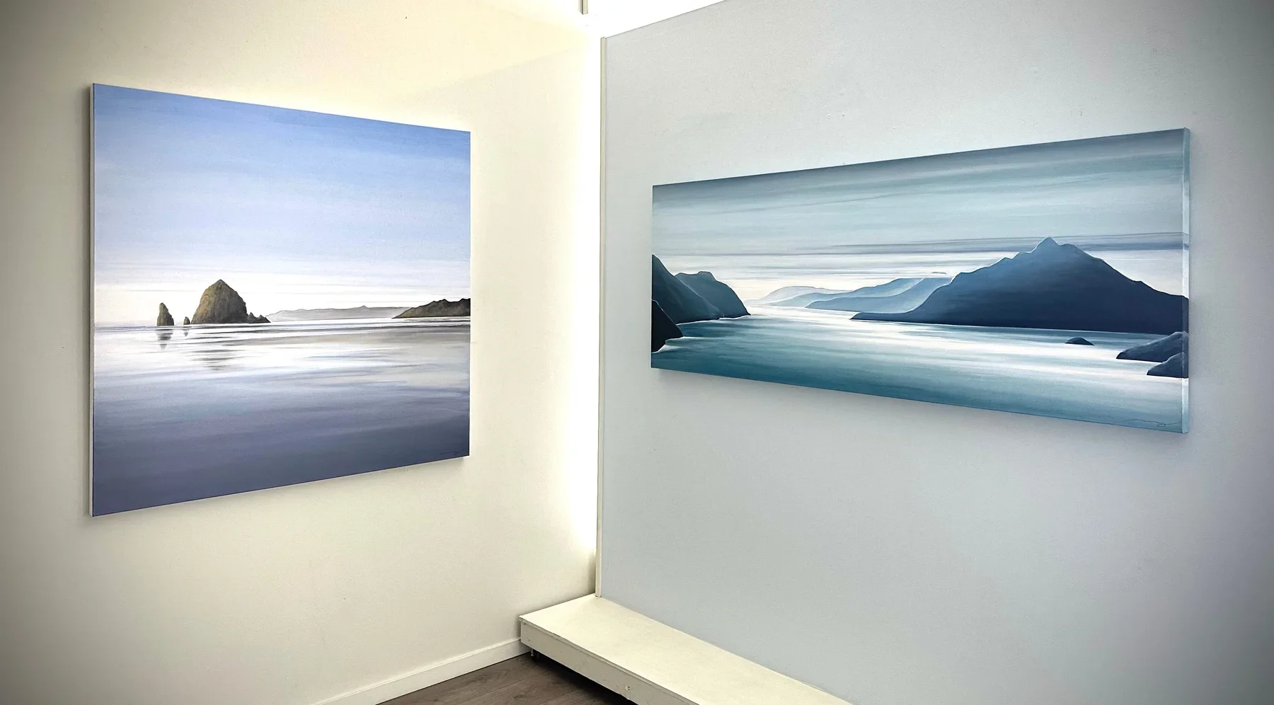Two landscape paintings of ocean and mountains hanging on white gallery walls.