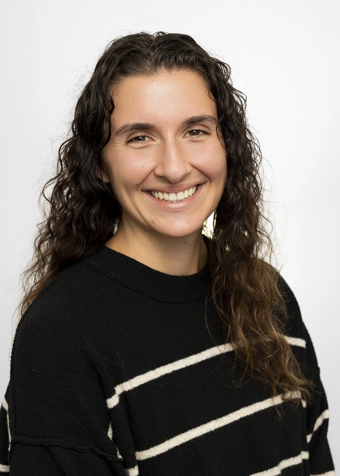 Portrait of a young woman with long, curly brown hair, smiling, wearing a black sweater with white horizontal stripes, against a plain white background.