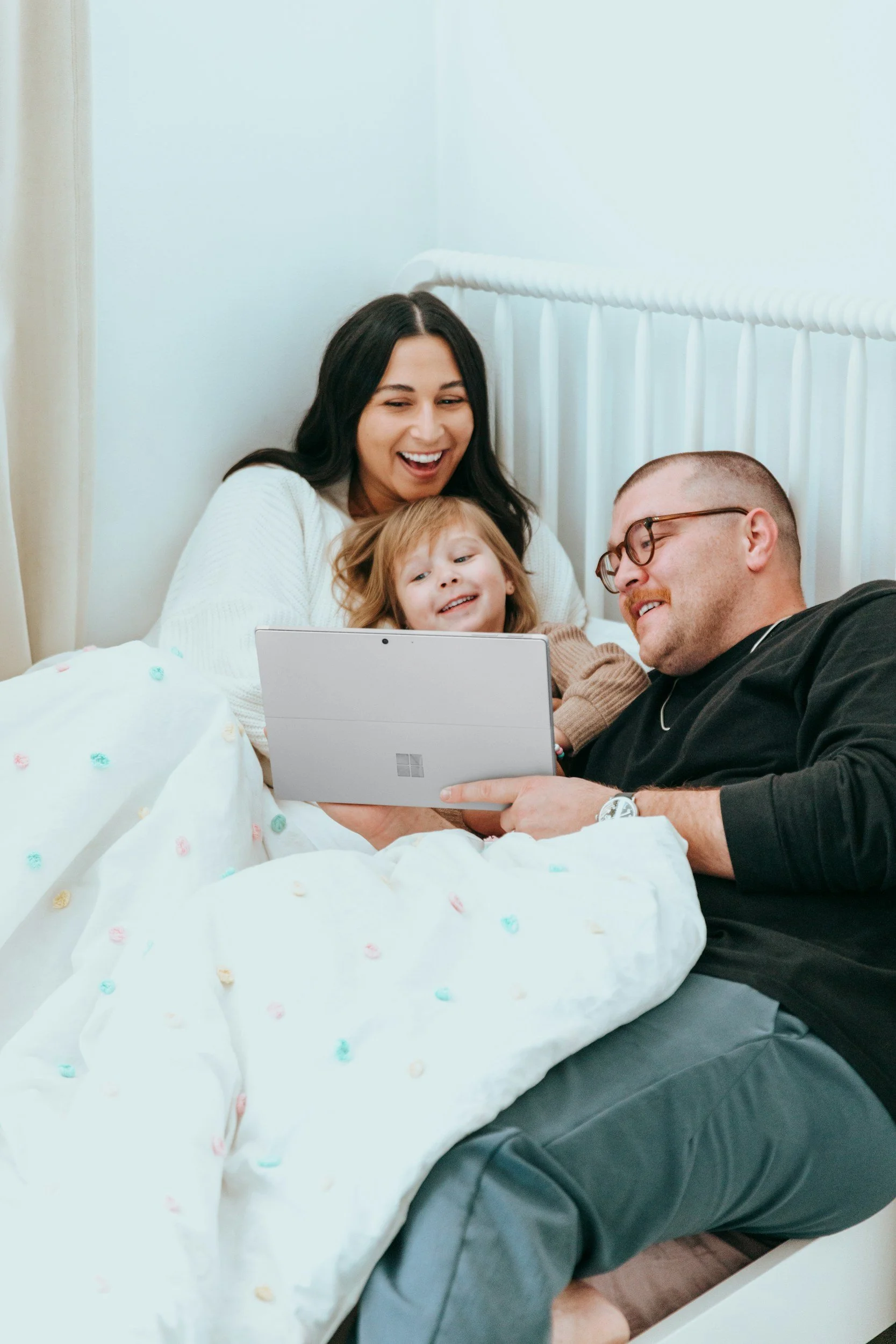 A family of three, a woman, a man, and a young girl, lying in bed together and looking at a tablet, smiling and enjoying their time.